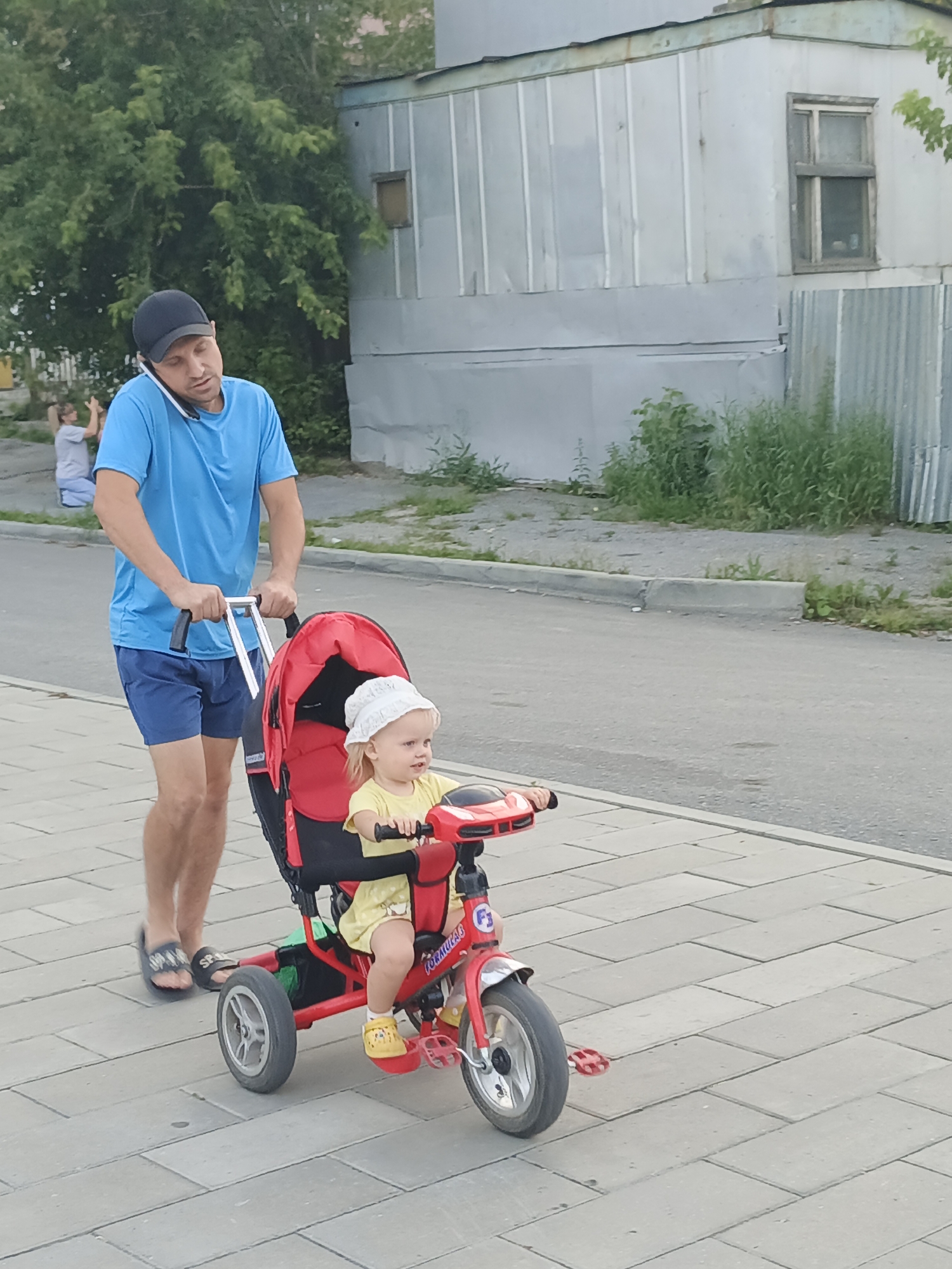 A man in a blue t-shirt and blue shorts is pushing a red stroller with a child in it. The man is holding a phone to his ear and is walking on a sidewalk made of light gray square pavers. The stroller has a black seat and a red canopy. The child is wearing a yellow t-shirt and a white hat. The child is holding onto the handlebars of the stroller. In the background, there is a metal building with a window. There is also a curb and a road. The scene is likely in Zlatoust, Russia.