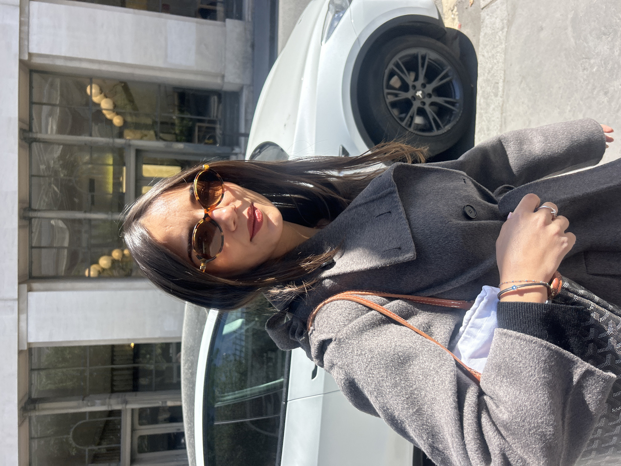 A medium shot shows a woman wearing sunglasses and a grey wool coat with a brown leather strap from a purse. She stands next to a white car with black rims in an urban environment. A building facade with windows is partially visible in the background.