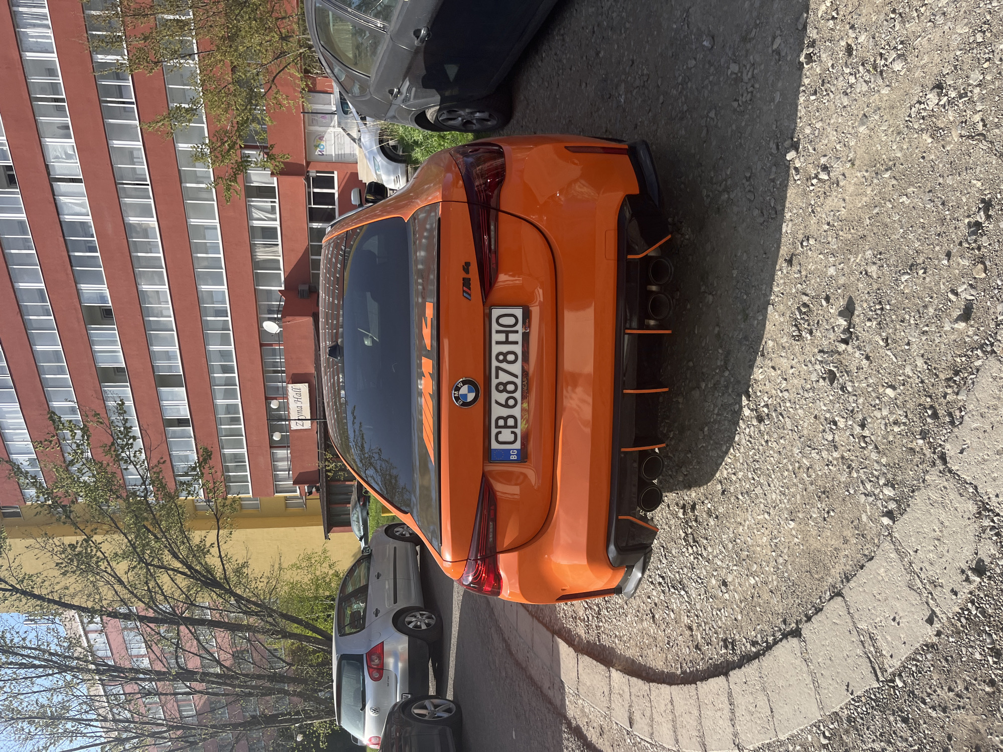 The image captures an orange BMW M4 parked on a gravel and concrete surface. The car's license plate reads "CB 6878 HO," indicating it is registered in Bulgaria. In the background, there is a tall building, with a sign that appears to read "Zoma Hall" written in Cyrillic. The building's color and architecture suggest a location in Sofia, Bulgaria. Other vehicles are parked nearby, adding to the urban street scene. The lighting suggests it is daytime.