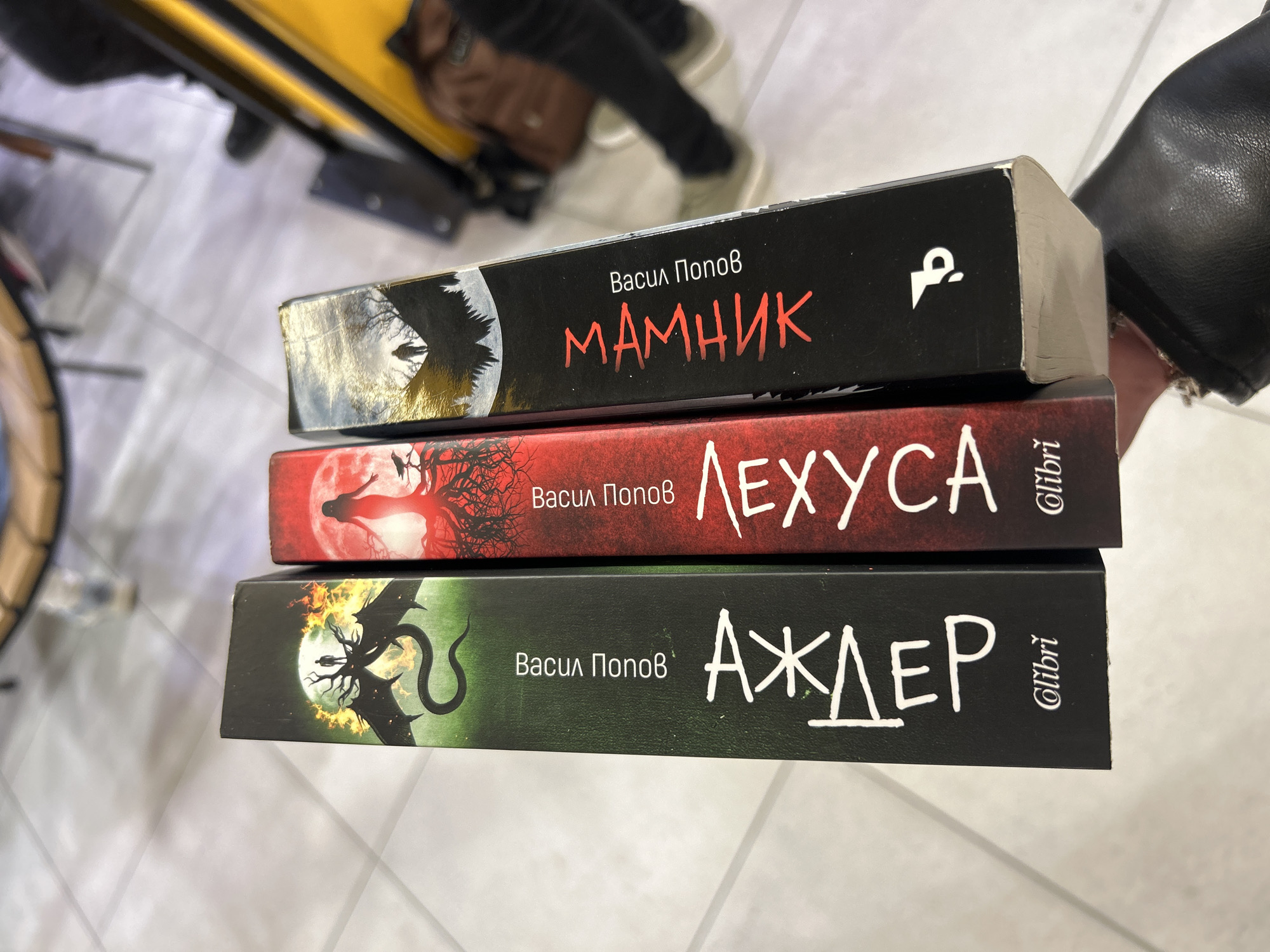 Three books with dark, fantasy-themed covers are stacked horizontally and held by a person in an indoor setting. The image is taken from a slightly elevated, oblique angle, with the books dominating the foreground. The background is blurred, showing parts of a person's leg and shoe, as well as an indistinct yellow object and a light-colored floor or surface, suggesting a casual environment like a bookstore or home in Plovdiv, Bulgaria.
The top book has a black spine featuring an illustration of a dark, winged creature or figure silhouetted against a full moon and bare trees. The author's name, "Васил Попов" (Vasil Popov), is printed in white above the title "МАМНИК" (Mamnik) in a stylized red font. A white, abstract logo is also visible on the right side of the spine.
The middle book has a deep red spine. Its illustration depicts a ghostly, ethereal figure and twisted, leafless trees against a large, glowing red full moon. The author "Васил Попов" is again in white, and the title "ЛЕХУСА" (Lehusa) is in a clean white font. The publisher's logo, "Olibri," is visible vertically on the right.
The bottom book features a dark green spine with an illustration of a black, dragon-like creature with spread wings and a serpentine tail, surrounded by fiery elements, against a full moon. Similar to the others, "Васил Попов" is printed in white, followed by the title "АЖДЕР" (Azhder) in a white, angular font. The "Olibri" logo is also present vertically on the right.
The visible text confirms Vasil Popov as the author for all three books, with titles Mamnik, Lehusa, and Azhder. The presence of the "Olibri" logo suggests these books are published by the Bulgarian publisher of the same name. The overall aesthetic of the book covers points towards genres of fantasy, horror, or dark fiction.
