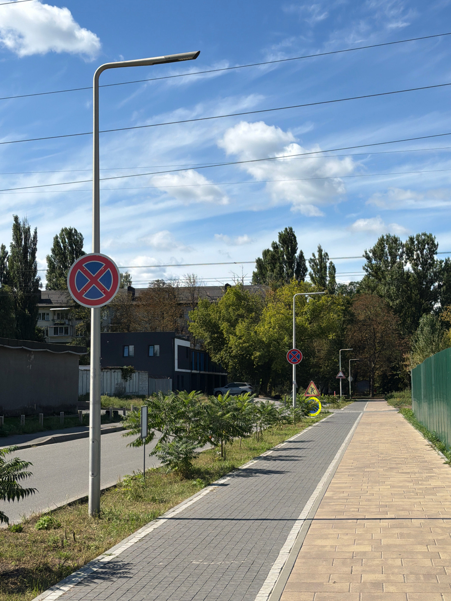 The image depicts a street scene in Kyiv, Ukraine, on a bright, partly cloudy day. In the foreground, a paved walkway with light brown and gray stone tiles runs alongside a road. A lamppost stands next to the walkway. A circular traffic sign with a red border and a blue "X" inside is visible on the lamppost. Further down the road, there are more lampposts and traffic signs, including a yellow triangular sign with a pedestrian symbol and a yellow circle. Trees line the street, with buildings and a green fence visible in the background. A silver car is parked on the side of the road. The sky is blue with scattered white clouds, and power lines stretch across the frame.