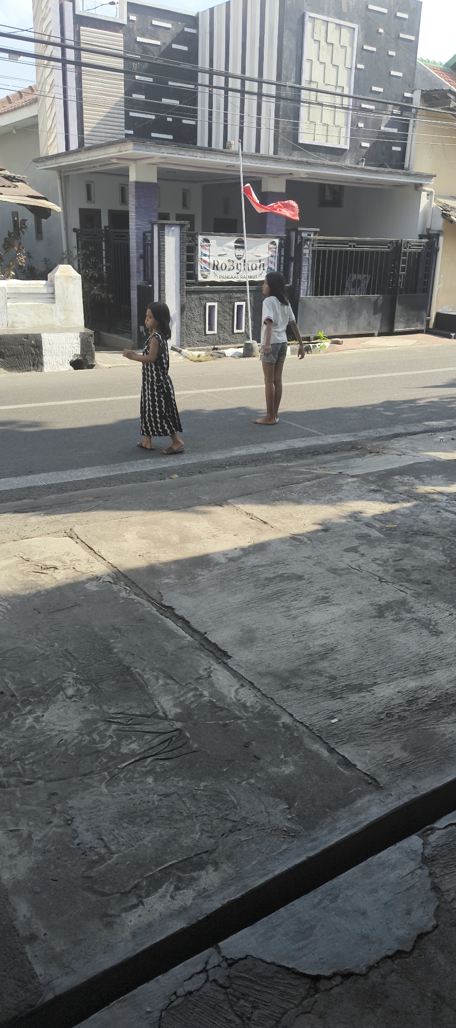 In Probolinggo, Indonesia, two young girls are seen on a street. The girl on the left is wearing a black and white patterned dress and appears to be walking. The girl on the right is wearing a white t-shirt and grey shorts, standing with her back to the camera. They are in front of a building with a modern facade featuring vertical black and white stripes. A red and white flag is flying from a pole in front of the building. A sign with the words "RoByhan" and "PANGKAS RAMBUT" (which means barbershop in Indonesian) is visible on the building. The street has white lines marking lanes, and a grey concrete sidewalk is visible in the foreground, showing signs of wear and tear. The scene is illuminated by sunlight, casting shadows on the ground.