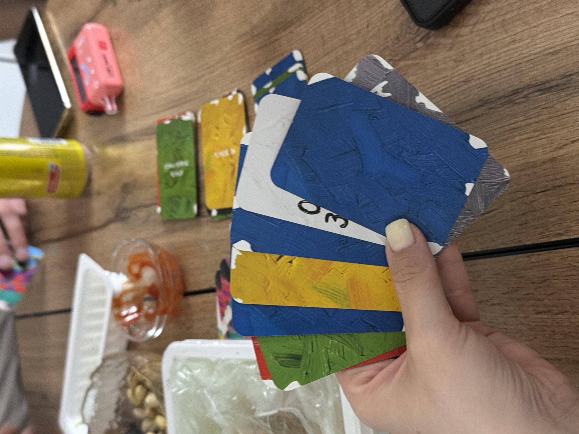 A top-down shot depicts a hand holding a stack of colorful, painted cards on a wooden surface. Adjacent to the cards are a disposable food container filled with clear plastic-wrapped contents, a small bowl of orange sauce, a yellow juice box, a pink vape pen, and a black mobile phone. A second individual's hand is partially visible on the left, holding a multicolored card. The scene appears to be indoors, set on a wooden table. The location is not discernible as Reshetnikovo, Russia.