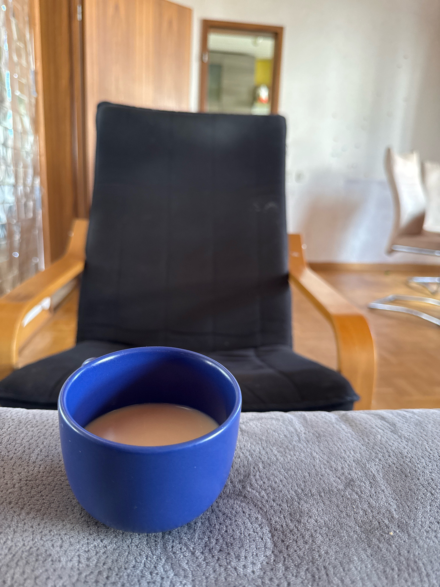 In a domestic indoor setting, likely a living room in Überherrn, Germany, a blue ceramic mug filled with a light-colored liquid, possibly coffee with milk, rests on a gray textured fabric surface. In the background, a dark fabric recliner chair with wooden armrests is visible. Further in the background, a wooden cabinet and a mirror are present. The lighting suggests it is daytime, possibly with natural light filtering in. There are no people or animals in the scene.