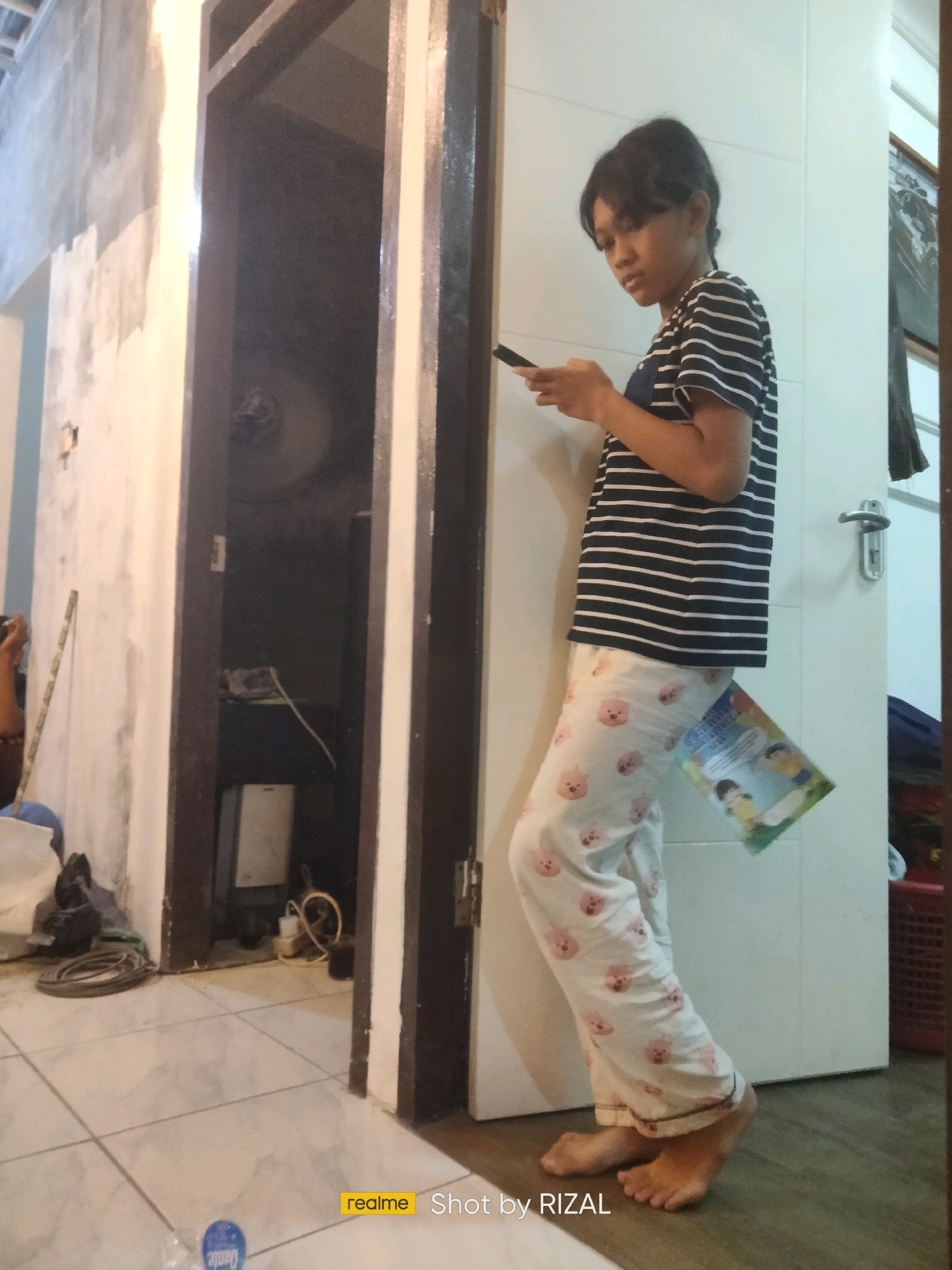 A young person stands near a doorway, looking down at a smartphone in their hands. They are wearing a striped t-shirt and pajamas with a cartoon bear pattern. A book is tucked into the waistband of their pajamas. The doorway opens into a dimly lit room with a fan and other objects. The floor is tiled, and a water bottle is visible near the bottom of the frame. The image appears to have been taken in a residential setting in Serdang Kulon, Indonesia, as indicated by the "realme Shot by RIZAL" watermark.