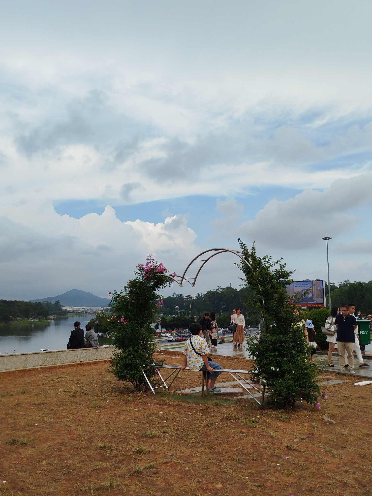 The image captures a scene at Xuan Huong Lake in Da Lat, Vietnam. A decorative metal arch with foliage, including pink flowers, frames a partial view of the lake and surrounding landscape. Several people are present in the scene: some are sitting on a bench integrated into the arch structure, while others are walking along the adjacent walkway. The ground around the arch is reddish-brown. In the background, the lake stretches towards a distant mountain, partially obscured by clouds. The sky is overcast, with patches of blue visible amidst the cloud cover. A streetlight and a billboard can be seen further in the background.