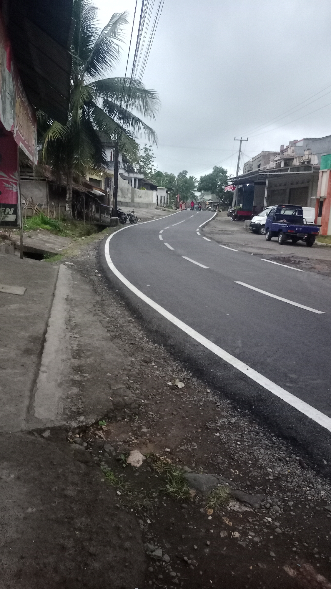 In Cogreg, Indonesia, a two-lane asphalt road curves gently to the right. A white solid line marks the edge of the road, while dashed white lines divide the lanes. To the left of the road, a concrete sidewalk is bordered by a drainage ditch and vegetation. Further left, a palm tree stands next to a low-slung structure with a corrugated metal roof. On the right side of the road, several buildings line the street, some with awnings and open storefronts. A dark blue pickup truck and a white van are parked in front of one of the buildings. In the distance, a few people are walking along the road. The sky is overcast, suggesting a cloudy day. The overall scene depicts a typical village road in a tropical climate, with a mix of residential and commercial structures.