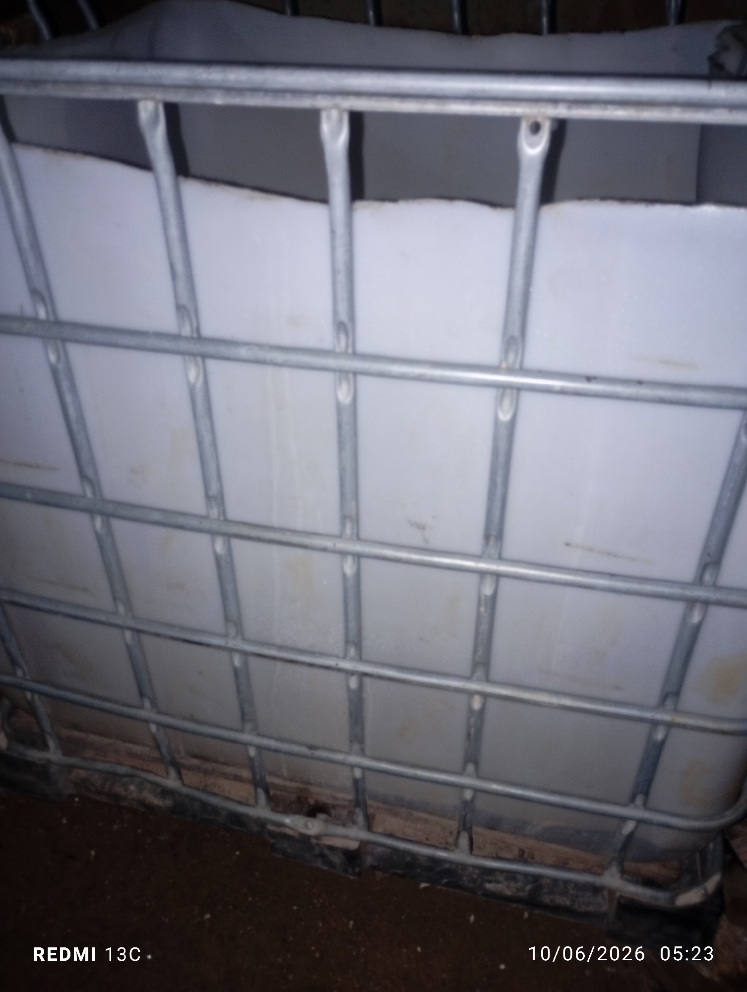 The image shows a close-up view of a large, white, plastic container encased in a metal cage. The container appears to be an Intermediate Bulk Container (IBC), commonly used for storing and transporting liquids. The metal cage is made of galvanized steel bars, forming a grid pattern around the container. The white plastic is smooth and shows some slight discoloration or dirt marks in places. The bottom of the container rests on a black plastic pallet with wooden supports. The background is dark and out of focus, suggesting an indoor or dimly lit outdoor environment. The lower portion of the image displays a timestamp and model information, indicating "REDMI 13C" and "10/06/2026 05:23". No people are visible in the image. The scene is static and appears to be a simple object observation. There is no indication of the specific location within Jos, Nigeria, other than the presence of the timestamp.