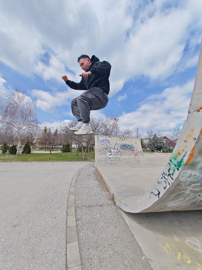 The image captures a dynamic outdoor scene at a concrete skatepark, located within the Youth Park (Младежки парк) in Shumen, Bulgaria. A young adult male is suspended in mid-air, positioned centrally in the frame. He is wearing a black hooded sweatshirt, dark grey sweatpants, and light-colored athletic shoes. Both hands are gripping a black handlebar, indicating an ongoing trick or jump performed with a scooter, which is not visible. His knees are bent upwards, and his arms are slightly flexed.
To the right, a large, worn concrete quarter pipe or ramp rises sharply, displaying extensive multi-colored graffiti in black, white, green, orange, and yellow hues. Another concrete skatepark feature, a quarter pipe, is visible in the midground, also marked with graffiti. The immediate foreground consists of a light-colored paved surface, separated by a curved grey stone curb from a narrow strip of gravel.
The background presents a park-like setting featuring green grass and numerous deciduous trees, most of which are bare or sparsely leaved, suggesting late winter or early spring. Scattered evergreen shrubs are also present. Distant urban buildings and streetlights are faintly visible on the horizon. The sky is partially cloudy with patches of blue.