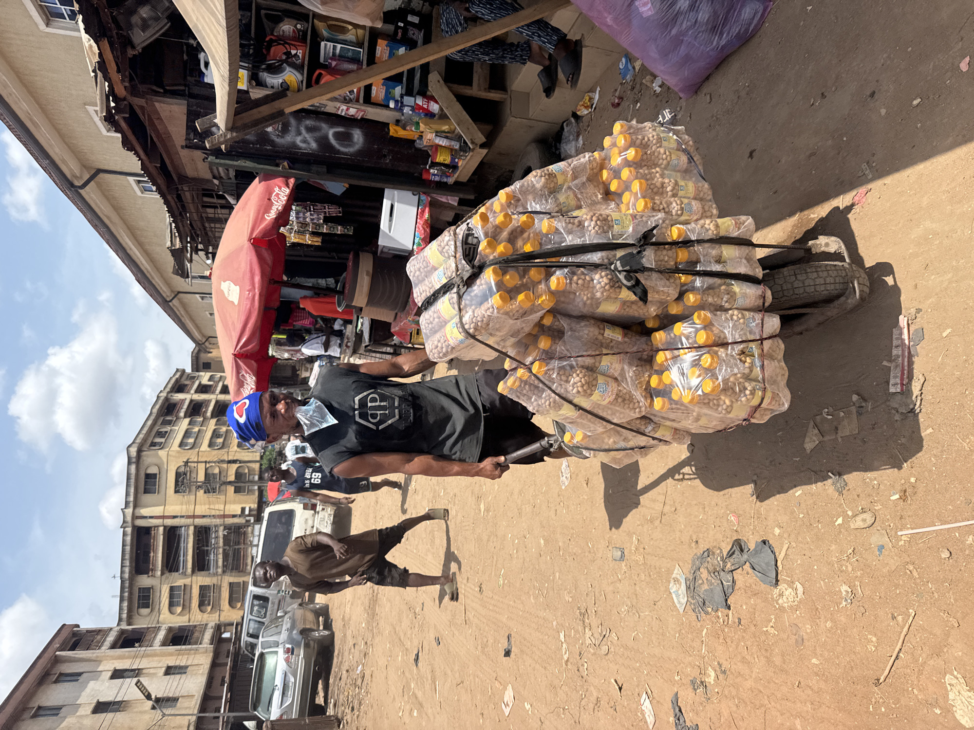 This image captures a bustling street scene in what appears to be an urban market under bright, sunny daylight. The ground is an unpaved, dusty light brown, littered with various debris including plastic bags and papers.
The central figure is a man, wearing a black t-shirt with a white logo (possibly "PP" or "DP"), dark trousers, and a blue cap adorned with a white heart. He has a clear plastic bag covering his mouth, serving as a makeshift face covering. He is actively pushing or pulling a single-wheeled cart heavily loaded with clear plastic bottles, each filled with light-colored, small, round snacks, likely nuts. These bottles are securely bundled onto the cart with black straps.
To the left of the main subject, a series of multi-story buildings with numerous windows line the street, suggesting a densely populated area. Several vehicles, including cars and a white van, are visible parked or moving further down the road. On the right side of the frame, there is a market stall or shop operating under a prominent red umbrella bearing the "Coca-Cola" logo. This stall displays various packaged goods. Another man, wearing a brown t-shirt and shorts, is visible walking in the mid-ground.
The clear blue sky with scattered clouds and distinct shadows cast by the subjects and objects indicate a sunny day, likely during mid-morning or afternoon. The overall scene depicts a vibrant commercial environment characteristic of a market in Onitsha, Nigeria, conveying a sense of everyday activity and commerce. Visible text includes "Coca-Cola" on the umbrella and a part of a logo on the central figure's shirt.