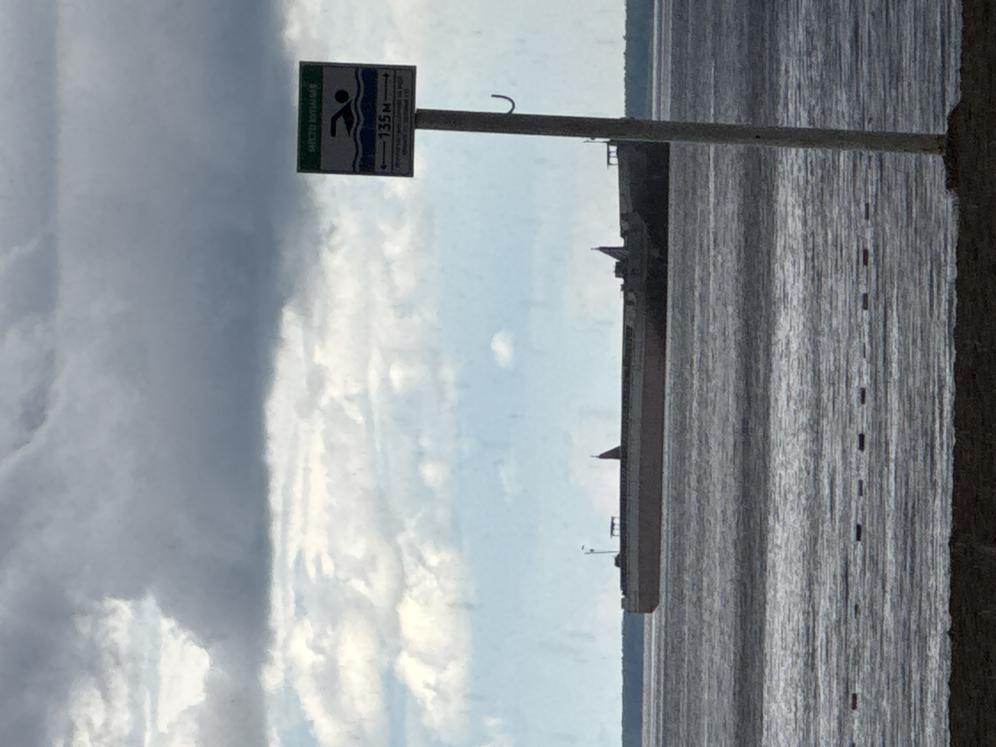 This media file captures an outdoor scene in Chaikovsky, Russia, depicting a body of water under a partly cloudy sky. The image is oriented vertically but should be viewed horizontally for correct interpretation.
In the foreground, a sign on a pole stands on what appears to be a bank or shoreline. The sign, green and blue, displays a pictogram of a swimmer and the Russian text "МЕСТО КУПАНИЯ," which translates to "SWIMMING AREA." An arrow points to the left with the number "135 М," indicating the swimming area is 135 meters in that direction. Smaller, less legible text below this seems to read "Граница пляжа," meaning "Beach boundary."
The expanse of water, likely a river or lake, fills the middle ground, showing ripples and a somewhat dark, reflective surface. A line of small, dark, rectangular markers or buoys is visible in the water, extending into the distance. On the far horizon across the water, several structures, including a building with a distinctive spire or roofline, are silhouetted against the lighter sky. The time of day appears to be daytime, with visible patches of blue sky amidst the clouds. No people are present in the scene.