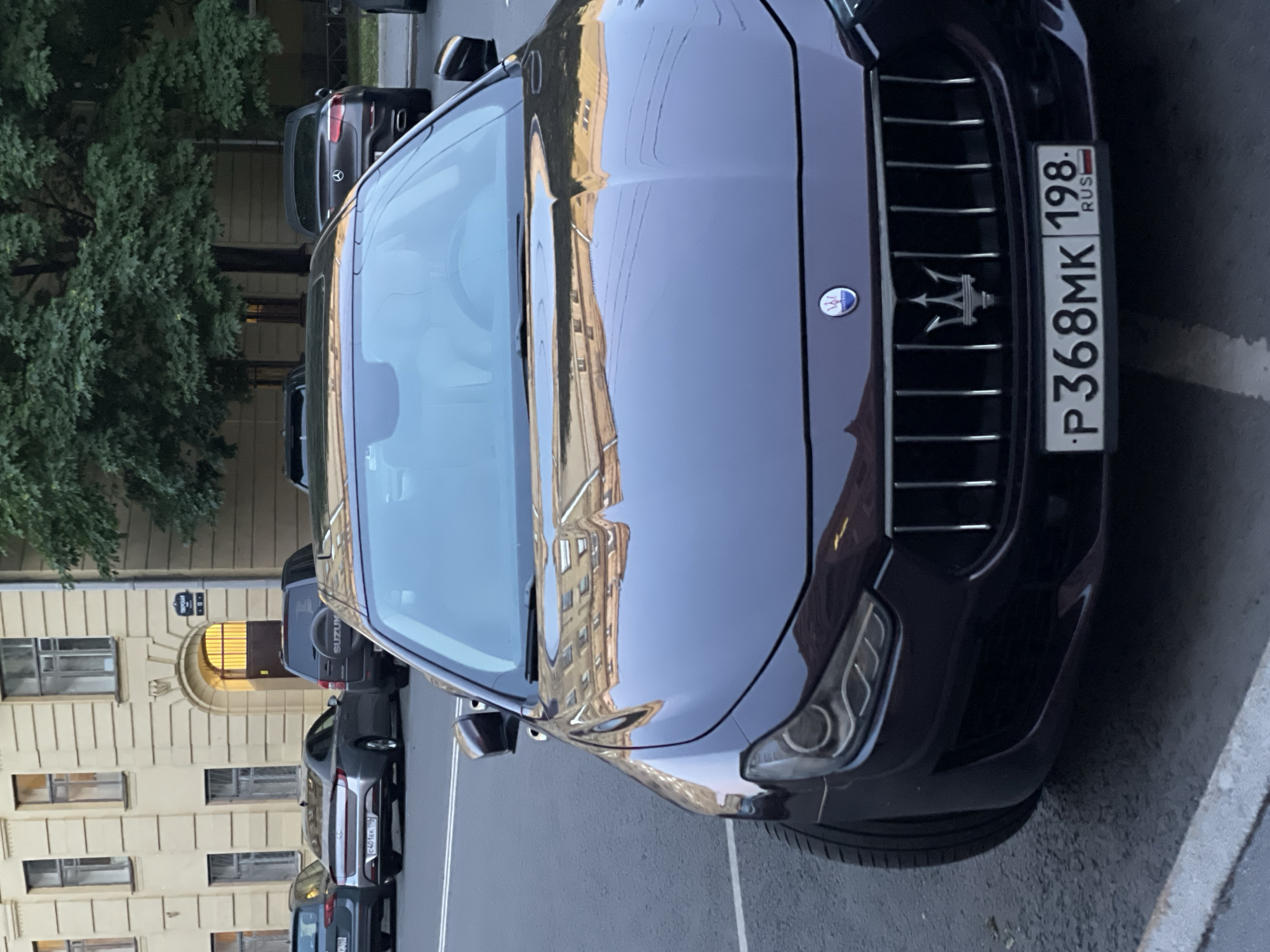 A dark-colored Maserati Ghibli is parked on a street in Saint Petersburg, Russia. The car is the primary subject, occupying most of the foreground and midground. It is facing forward, with its front grille and license plate visible. The license plate reads "P368MK 198 RUS". The car's emblem is a trident. In the background, a light-colored building with arched doorways and windows, and several trees with green foliage, are visible. Other cars are parked on the street in the background, including a dark Suzuki SUV and a dark Mercedes-Benz sedan. The lighting suggests it is daytime, likely late afternoon or early evening. The pavement is wet in some areas, indicating recent rain. The overall scene depicts a street view in a city setting.