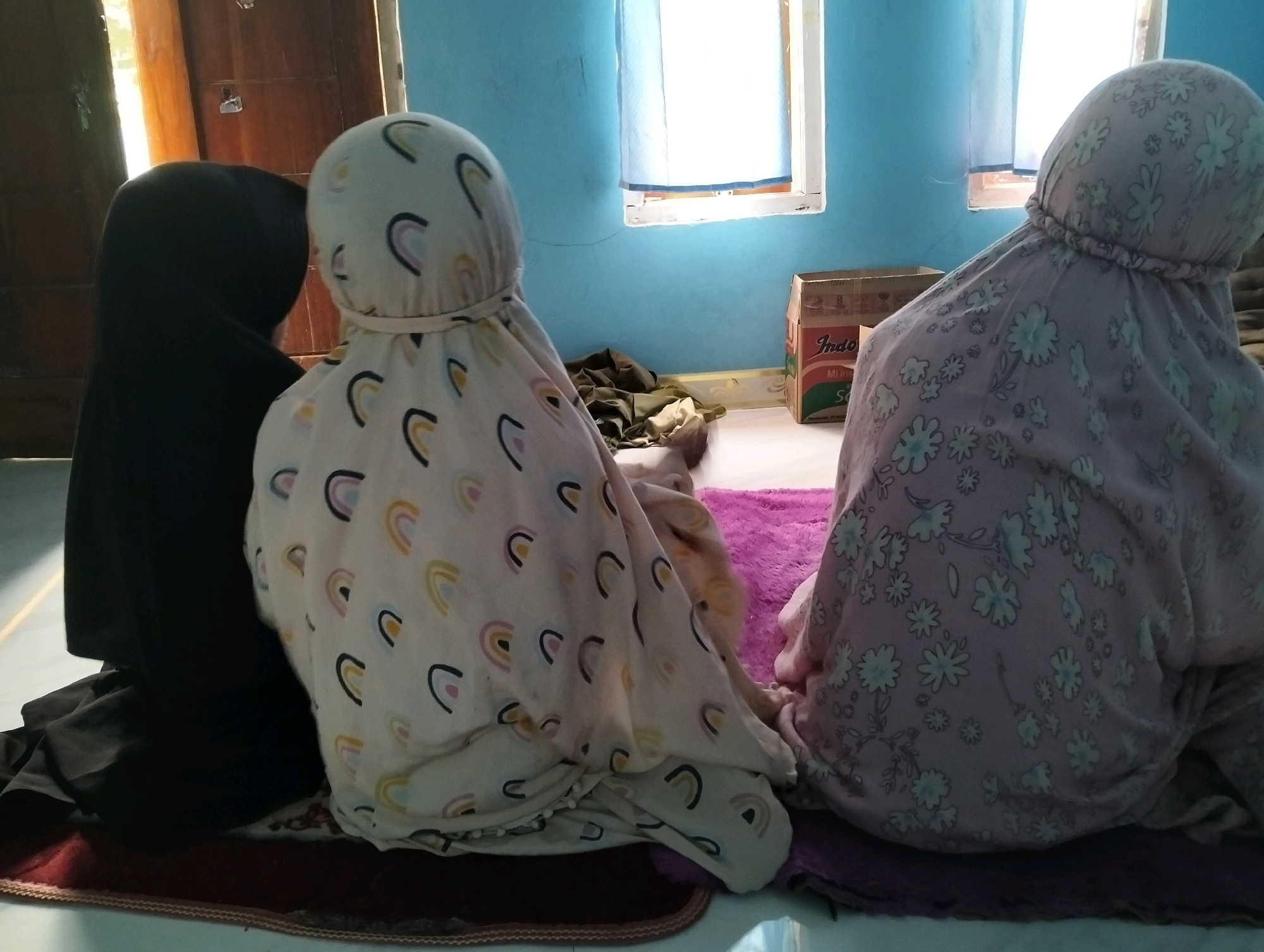 The image shows two individuals, likely children or young women, wearing headscarves and religious garments, sitting on prayer mats. One individual on the left is wearing a black headscarf and garment. The other two individuals are wearing patterned garments; one has a cream-colored headscarf and garment with rainbow motifs, and the other has a light pink headscarf and garment with floral patterns. They appear to be engaged in prayer or a similar religious activity within an indoor setting. The room has light blue walls, a window with white curtains, and a wooden door on the left. A cardboard box with "Indomie" branding is visible on the floor, along with a purple rug and other fabrics. The lighting suggests it might be daytime. This scene is typical of a domestic prayer space and could be located in Cileungsi, Indonesia, where such activities are common.