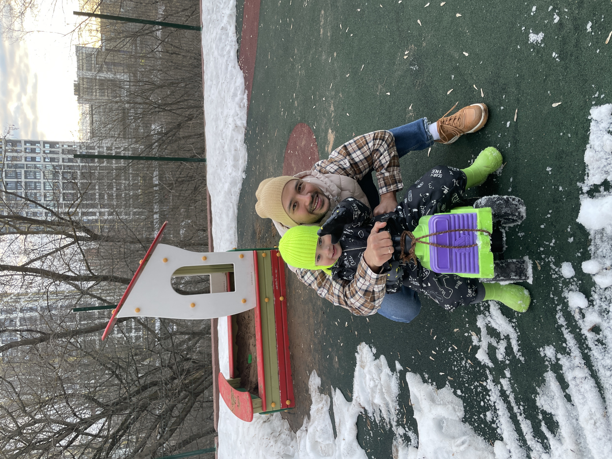 The image depicts a person and a child sitting on a sled in a snow-covered playground. The person wears a beige beanie, a plaid shirt, and blue jeans, and has brown boots on. The child wears a neon green beanie, a black jacket with white drawings, and neon green boots. They are holding a neon green and purple sled. Behind them, there is a sandbox with a red and white frame. The ground is partly covered with snow and partly with green and red playground surface. In the background, trees and a tall building can be seen. The location is Izhevsk, Russia.