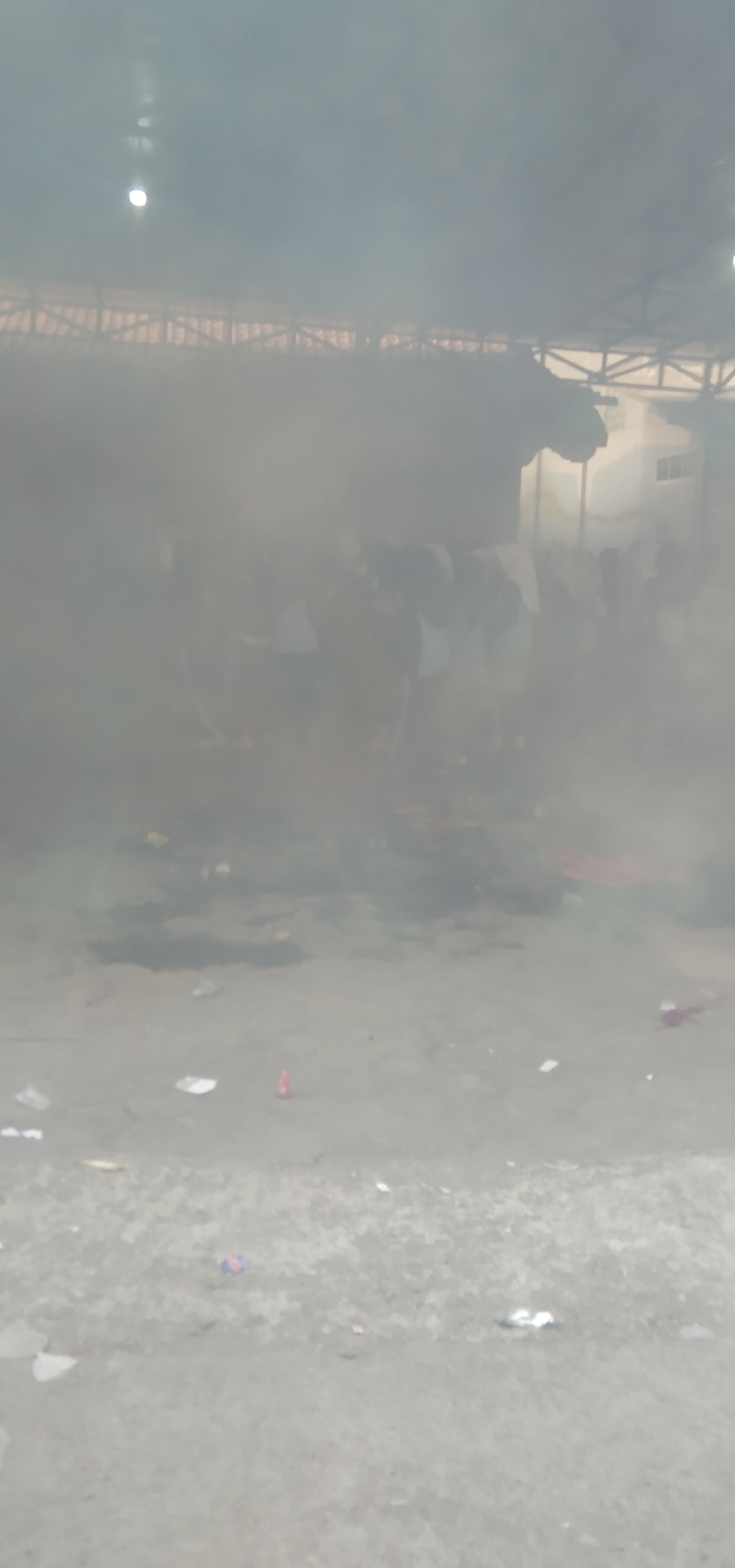 A smoky, somewhat blurry image depicts an outdoor scene under a partially covered structure, likely in Rahayu, Indonesia. The ground appears to be a dirt or concrete surface littered with various small objects, possibly trash or debris. In the background, a crowd of people is visible, mostly obscured by the heavy smoke. They are standing in rows and appear to be wearing dark clothing with some white elements. The overall impression is of an event or gathering taking place amidst dense smoke.