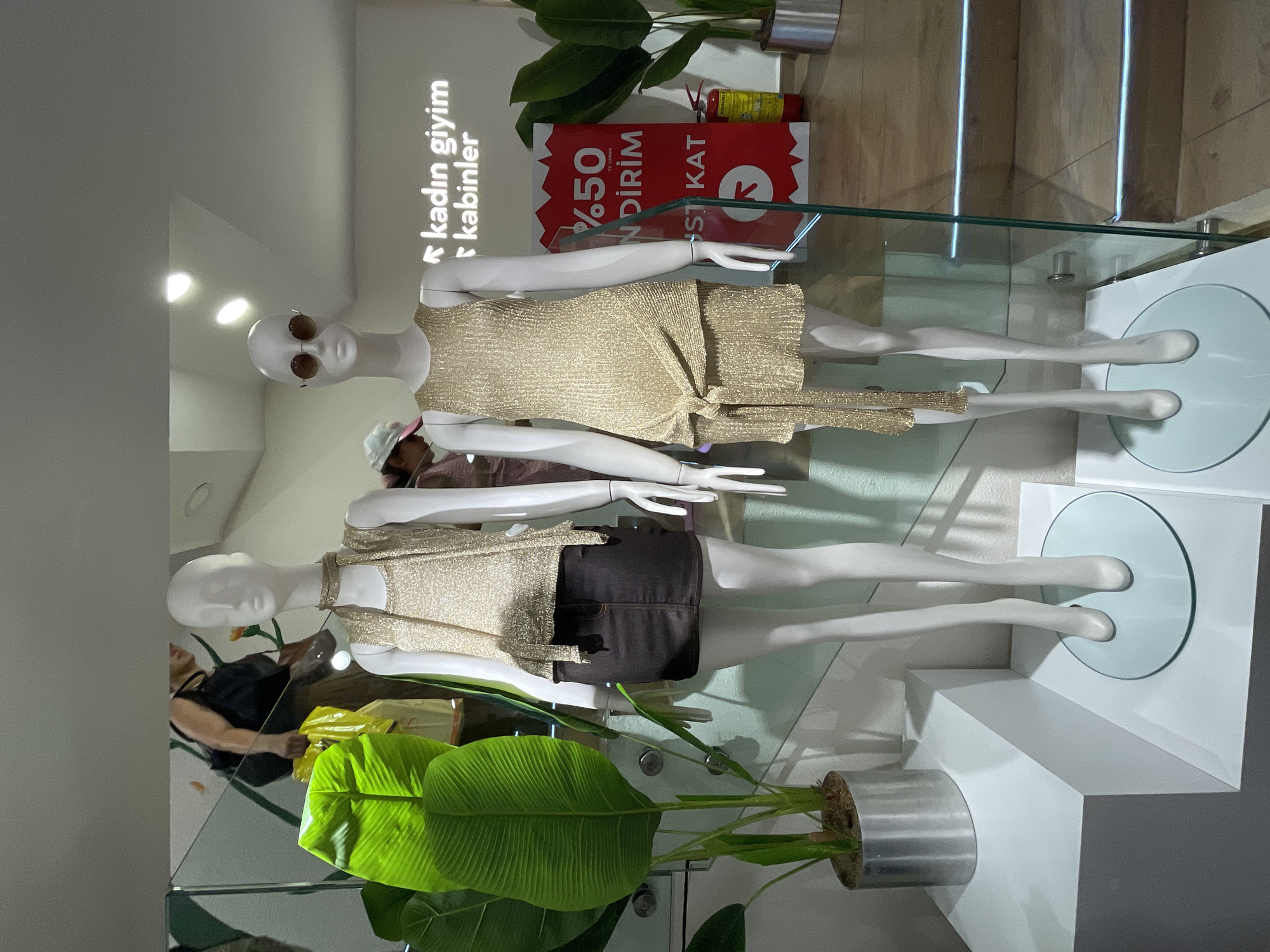 This image depicts a retail store window display in Kadıköy, Turkey. Two female mannequins are prominently featured, both wearing gold, sparkly outfits. The mannequin on the left is dressed in a gold sleeveless top with a fringed neckline and a dark brown skirt. The mannequin on the right wears a gold, sleeveless dress with a knot detail at the waist and sunglasses on its head. Behind the mannequins, a large, vibrant green leaf plant in a silver pot is visible. A sign with red and white text, partially obscured by a glass barrier, indicates a sale of "%50 İNDİRİM" (50% discount). Further in the background, stairs with illuminated edges lead upwards, and a fire extinguisher is mounted on the wall. A person, partially visible, is walking up the stairs carrying shopping bags. The overall environment suggests an indoor shopping space with modern display elements.