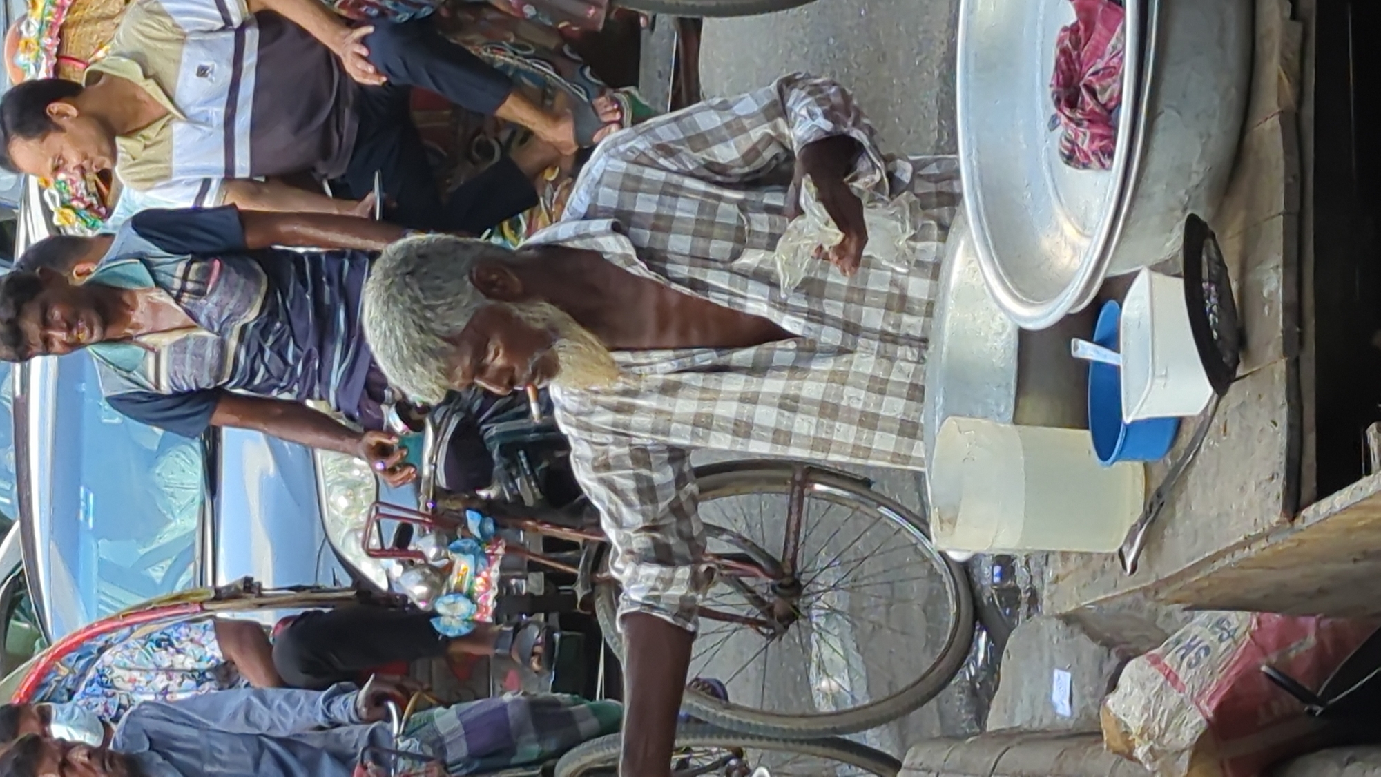 The image shows a street scene in Dhaka, Bangladesh. In the foreground, an elderly man with grey hair and beard, wearing a checkered shirt, is standing and holding a crumpled plastic bag. He appears to be smoking. To his left, a large metal basin and a blue bowl are visible on a wooden surface, along with a jug of water and a strainer. In the background, several men are seen in rickshaws and on foot, some wearing masks. The overall environment suggests a busy urban setting with traffic and pedestrians.