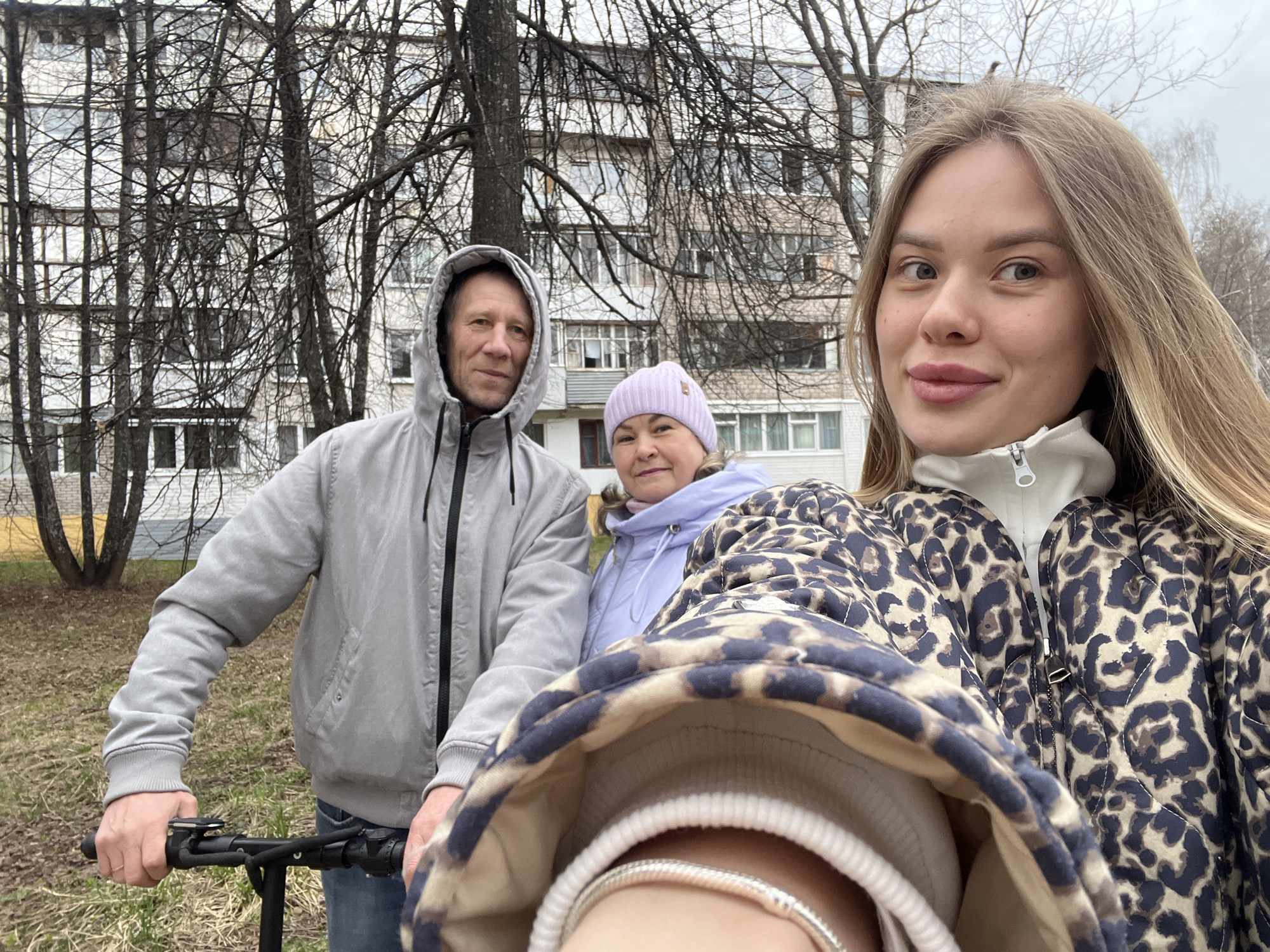 The image presents a selfie of a young woman with two older adults, possibly her parents, in an urban outdoor setting. The woman is in the foreground, wearing a brown and black animal print jacket over a white turtleneck sweater. A silver bracelet is visible on her wrist. To her left, a man wearing a grey hooded jacket is partially obscured by the woman's jacket; he is holding onto the handlebars of what appears to be an electric scooter. Behind them, an older woman wearing a lilac jacket and a pink knit hat stands. They are all looking toward the camera.
The backdrop consists of a multi-story residential building, typical of Soviet-era architecture, partially obscured by bare trees. The ground is covered with brown grass and some leaf litter, suggesting early spring or late autumn.
Based on the metadata from the image, it was taken in Chaikovsky, Perm Krai, Russia.