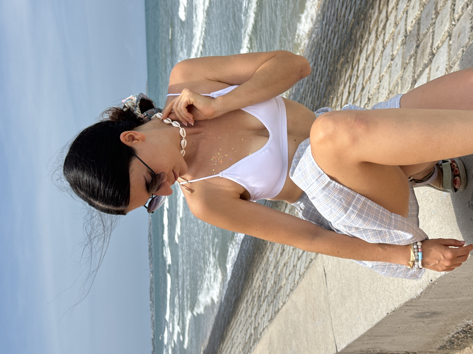 The image shows a young woman sitting on a concrete barrier overlooking the ocean in Mũi Né, Vietnam. She has dark hair pulled back in a bun with a patterned scrunchie and is wearing sunglasses, a white bikini top, and a light blue plaid skirt. A shell necklace adorns her neck, and she has glitter on her chest. She is also wearing several bracelets on her left wrist and sandals on her feet. The sea is visible in the background with gentle waves. The sky is clear and blue. The lighting suggests it is daytime.