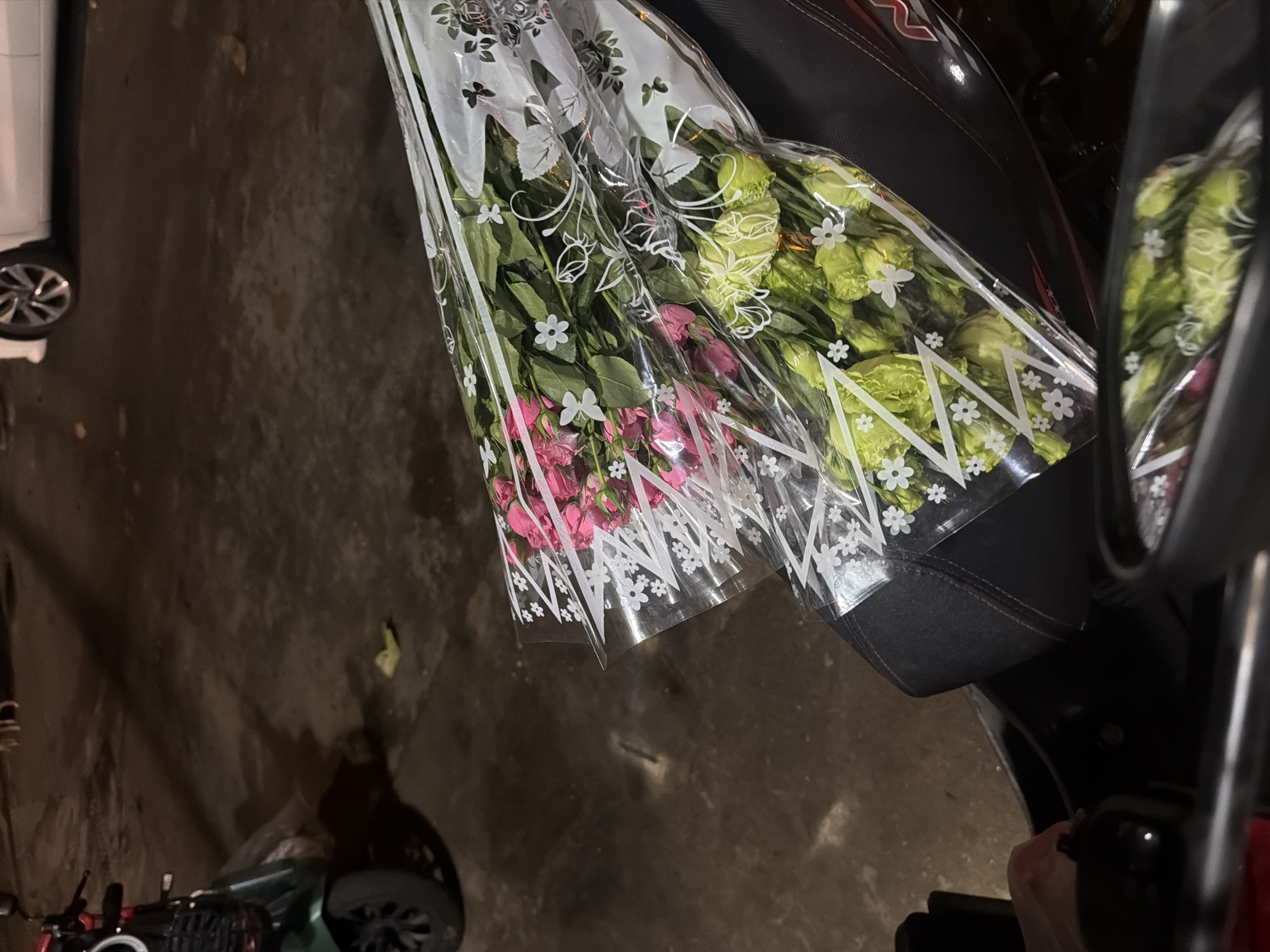 The image depicts two bouquets of flowers, one with pink roses and the other with light green flowers, wrapped in clear plastic with white floral patterns. They are placed on the seat of a dark-colored motorcycle. In the background, the ground appears to be asphalt or concrete, with some debris visible. A green scooter is parked to the left, and a white car is partially visible in the upper right corner. The lighting suggests it is nighttime or dusk. The scene appears to be an outdoor location, possibly a street or parking area in Nha Trang, Vietnam, given the context.