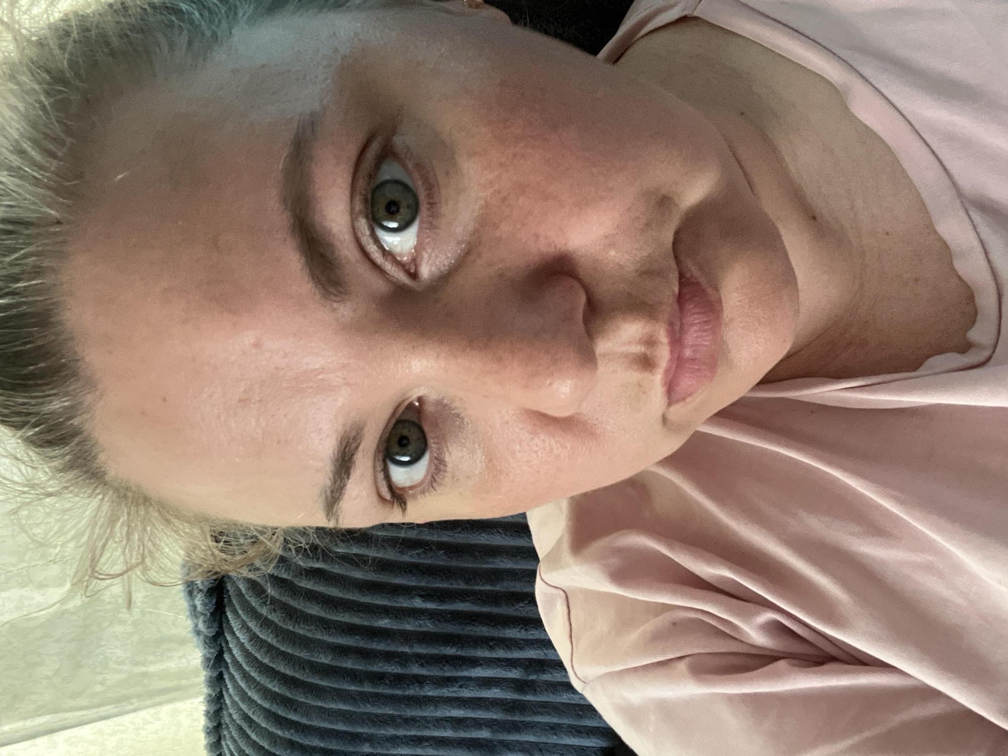 A close-up, medium shot shows a woman's face looking directly at the camera. She is sitting in front of a dark blue, corduroy-like textured surface, possibly a couch. The background is a blurry, light-colored curtain. The woman has blonde hair pulled back, and she is wearing a light pink t-shirt. Her eyes are green, and her expression is neutral. There is no indication of the location being in the city of Foky, Russia.