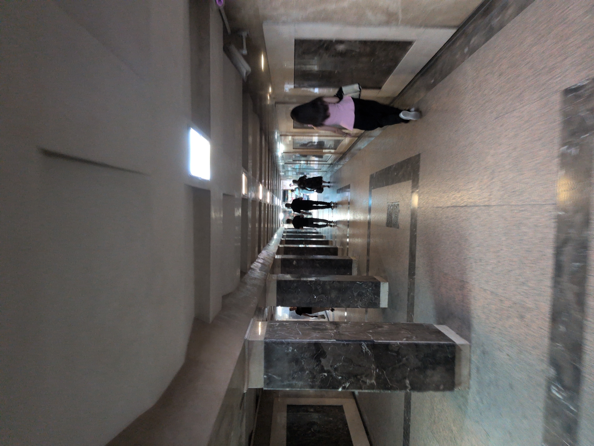 An interior shot of a pedestrian underpass shows a long, covered walkway with marble-clad pillars on either side. The floor is tiled with a mosaic pattern of beige and grey tiles, with darker grey bands at intervals. Several overhead fluorescent lights illuminate the space, with one particularly bright light panel visible towards the center of the walkway.
Several individuals are walking in both directions along the underpass. In the foreground, a woman with long dark hair is walking away from the camera, wearing a pink top and black pants. Further down the walkway, a group of three men are walking towards the camera. Another woman in a dark dress is visible in the distance. Security cameras are mounted on the wall on the right side of the underpass. The overall impression is one of a functional, urban transit space.