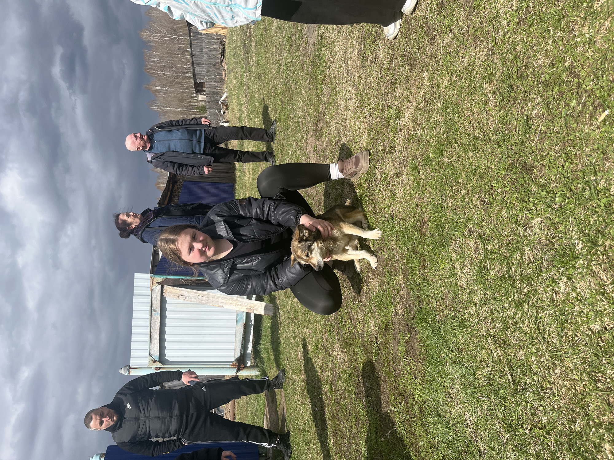 An outdoor scene captures several individuals on a grassy patch in what appears to be a rural setting in Буренка, Russia. In the foreground, a young woman is squatting, gently holding and petting a small to medium-sized dog with light brown and white fur. Her expression is calm, and the dog appears content.
To the left, a man in a black tracksuit stands, his jacket displaying a "NIKE" logo and his pants showing "Adidas" stripes. In the middle ground, an older bald man stands observing. Another person is partially visible behind the young woman, and legs/feet of other individuals are seen at the top and bottom edges of the frame.
The background features various structures, including fences made of light-colored corrugated metal sheets, some blue sheeting, and rustic wooden buildings. Beyond these, bare trees line the horizon. The sky is notably overcast and dark with heavy clouds on the left side, suggesting a stormy weather front, while other areas of the scene are lit by bright sunlight, casting visible shadows. A white cigarette butt is discernible on the grass in the lower right foreground. The overall impression is a casual daytime gathering, possibly in a yard or open field, with a focus on interaction with the dog.