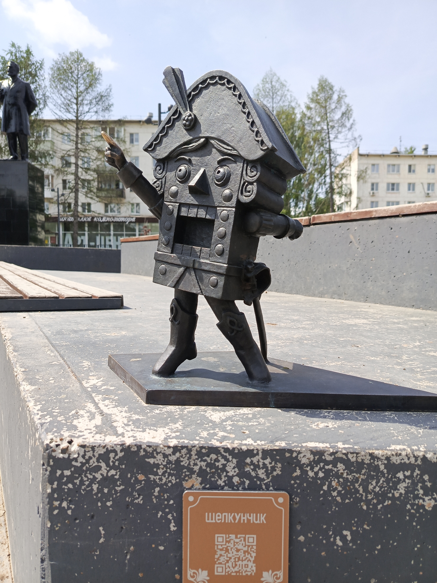 The image depicts a metal sculpture of a Nutcracker, standing on a concrete base. The sculpture has a rectangular body with decorative elements resembling buttons, a head shaped like a building facade with a tall hat, and outstretched arms, one pointing upwards. The sculpture stands on a metal platform, which sits atop a concrete base that is weathered and has a sign attached to it with the name "ШЕЛКУНЧИК" and a QR code. In the background, there is a wooden bench, another statue on a black pedestal, buildings, and trees. The image appears to be taken outdoors on a bright day. The location is likely in the city of Chaikovsky, Russia, as indicated by the Russian text on the sign.