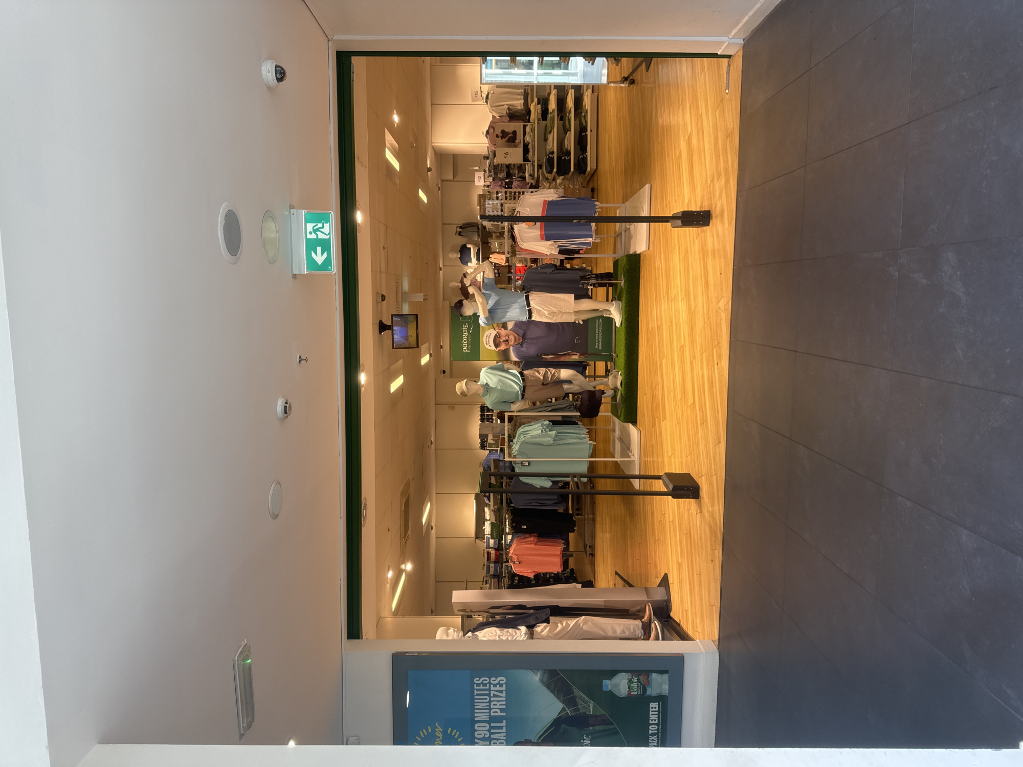 An exterior view of a retail store entrance reveals a well-lit interior filled with merchandise and mannequins. The store appears to be a clothing retailer, possibly specializing in golf attire, given the presence of mannequins dressed in golf apparel and a display featuring golf-themed imagery.
The entrance is framed by a dark green border, and the floor inside is a light-colored wood, contrasting with the dark grey tiled flooring in the foreground. A security gate or sensor is visible in the middle of the entranceway.
On the left side, a large poster displays a promotional message with a picture of a bottle of Volvic water and text mentioning "90 Minutes" and "Ball Prizes." Two mannequins stand near the entrance, one dressed in a suit and the other in casual wear.
Inside the store, clothing racks are filled with various garments, predominantly polo shirts and t-shirts in shades of blue, green, and orange. Several mannequins are positioned throughout the store, showcasing different outfits. In the background, shelves are stocked with folded clothing.
Above the entrance, a green illuminated sign with an arrow and a running figure indicates an emergency exit. Other ceiling-mounted fixtures include lighting and what appear to be smoke detectors or surveillance cameras. The overall impression is that of a typical retail environment.