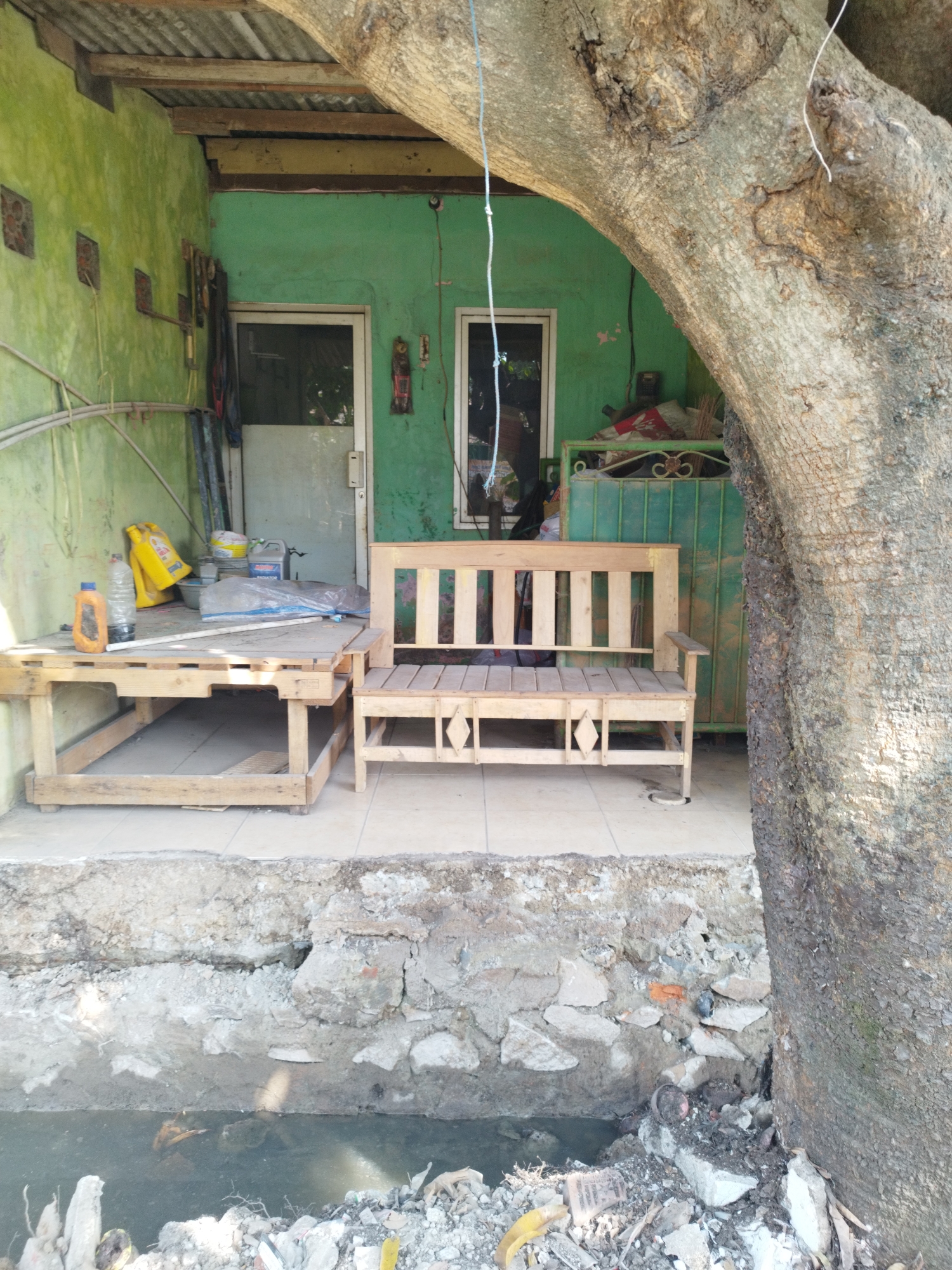 The image depicts an outdoor scene in what appears to be a residential or workshop area in Karawang, Indonesia. The foreground shows a section of rough, uneven ground with concrete debris and a small, murky body of water. A large, textured tree trunk dominates the right side of the frame, its bark rough and dark.
Behind the tree, a wooden bench with a slatted seat and back is positioned on a tiled patio. To the left of the bench is a simple wooden table, also with a slatted surface, which holds a few items including two yellow plastic containers, a clear plastic bottle with an orange cap, and a large, clear plastic bag. The wall behind the table and bench is painted a vibrant green. There is a doorway with a white frame and glass to the left, and another doorway, partially obscured by the tree, to the right. A corrugated metal roof structure is visible above the green wall. The overall impression is one of an unkempt, utilitarian outdoor space.