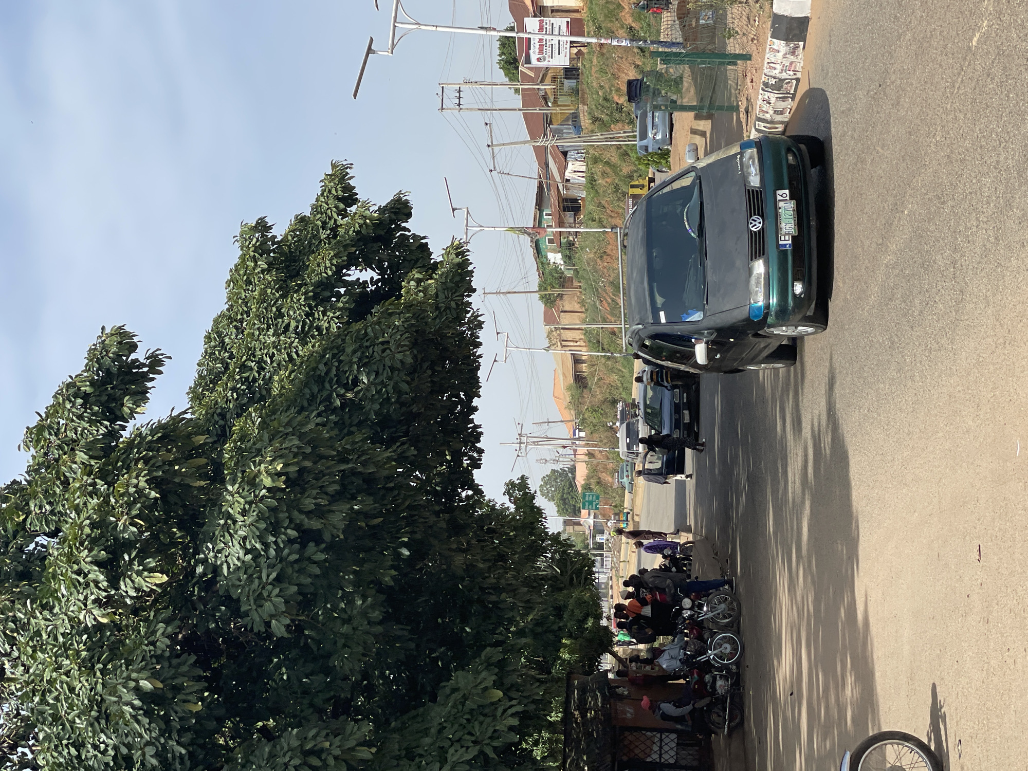 The image shows a street scene in Bukuru, Nigeria. A green Volkswagen car is prominently positioned on the right side of the road, partially obscuring other vehicles further down the street. Several people are gathered near parked motorcycles on the left side of the road. Overhead electrical wires and light poles stretch along the street, and buildings are visible in the background. A large tree dominates the left foreground of the image. The asphalt road appears well-maintained. The license plate of the Volkswagen is partially visible, with the characters "E-JGH 127D" legible.