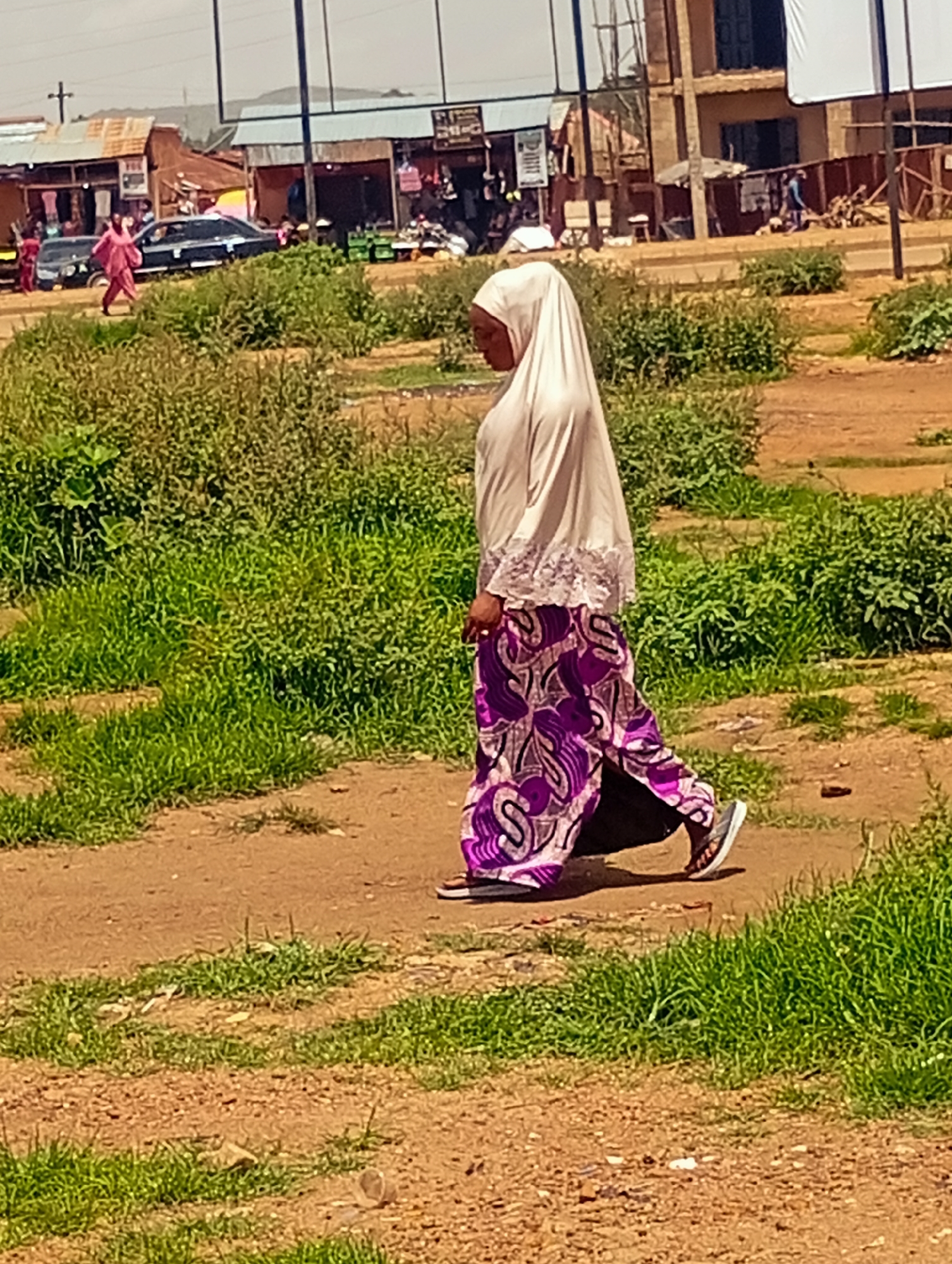 A woman walks through a dusty, sunlit area with a mix of grass and dirt. She is wearing a light-colored hijab and a long, flowing skirt with a vibrant purple and white pattern. Her gait suggests she is walking with purpose.
In the background, a collection of buildings lines a street. These structures appear to be a mix of shops and residential buildings, with some having corrugated metal roofs and others more substantial construction. Various goods are visible outside some of the shops, suggesting a marketplace or commercial area. A few cars are parked or driving on the road, including a dark sedan and a smaller red car. Other figures, including children, are visible near the buildings.
The scene is bright, indicating it is likely daytime, possibly mid-morning or afternoon, with clear weather. The overall setting appears to be a busy, somewhat informal urban or semi-urban environment in Bukuru, Nigeria. The environment suggests a typical day with people going about their activities. No specific text is clearly legible in the image.