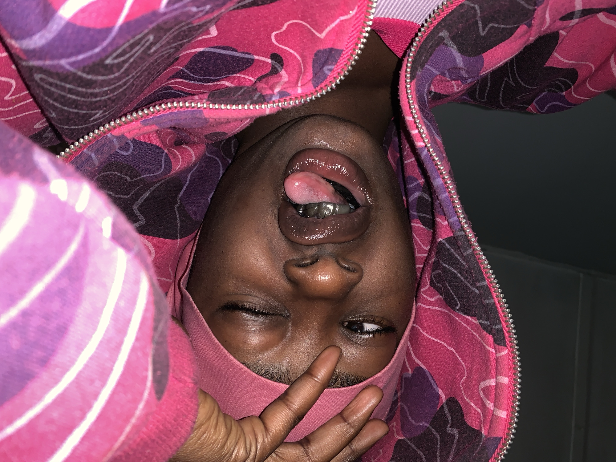A person with dark skin is upside down, wearing a pink head covering. One eye is open and one is closed. The person is sticking their tongue out and has silver grills on their bottom teeth. A hand is near the person's face. The person is wearing a pink and purple patterned jacket with a silver zipper. A gray wall is visible in the background. There is no indication the image was taken in Rafin Par, Nigeria.