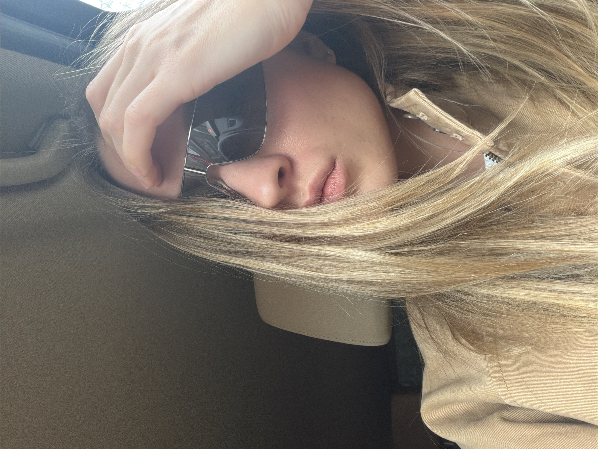 This is a close-up, rotated portrait of a person with long, light blonde hair, captured from a sideways perspective. The individual, likely a woman, is seen with a hand resting over their forehead and temple, partially obscuring the upper face. Large, reflective sunglasses cover one eye and part of the nose, while the other side of the face, revealing the nose and slightly parted lips, is visible amidst the hair.
The person is wearing a light beige or tan jacket with a visible zipper detail on the right side of the frame. The setting appears to be the interior of a vehicle, suggested by the light-colored headliner and a grab handle visible in the upper left. Another light-colored, possibly leather, interior element is seen towards the bottom, partially covered by the person's hair. Bright light, indicative of daylight, filters in from what appears to be a window in the upper background.
The activity depicted suggests the person is resting or relaxing, possibly while traveling in a car. The overall mood is one of introspection or casual repose. There is no visible text in the image. The provided location context for this image is Moscow, Russia.