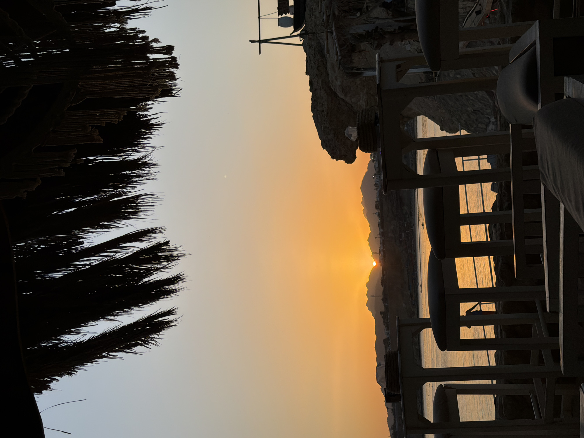 A silhouette of a thatched roof overhangs a serene sunset scene at Naama Bay Beach in Sharm El Sheikh, Egypt. The sun, a brilliant orange orb, dips below a distant mountain range, casting a warm glow across the calm sea. In the foreground, the dark outlines of outdoor seating, including tables and stools, are visible, hinting at a beachfront bar or restaurant. The water reflects the vibrant sky, and a few small boats are scattered on the surface. The rocky, natural landscape of the bay is visible in the background, with some structures and vegetation adding to the atmosphere. The overall impression is one of tranquil beauty and a perfect evening setting.
