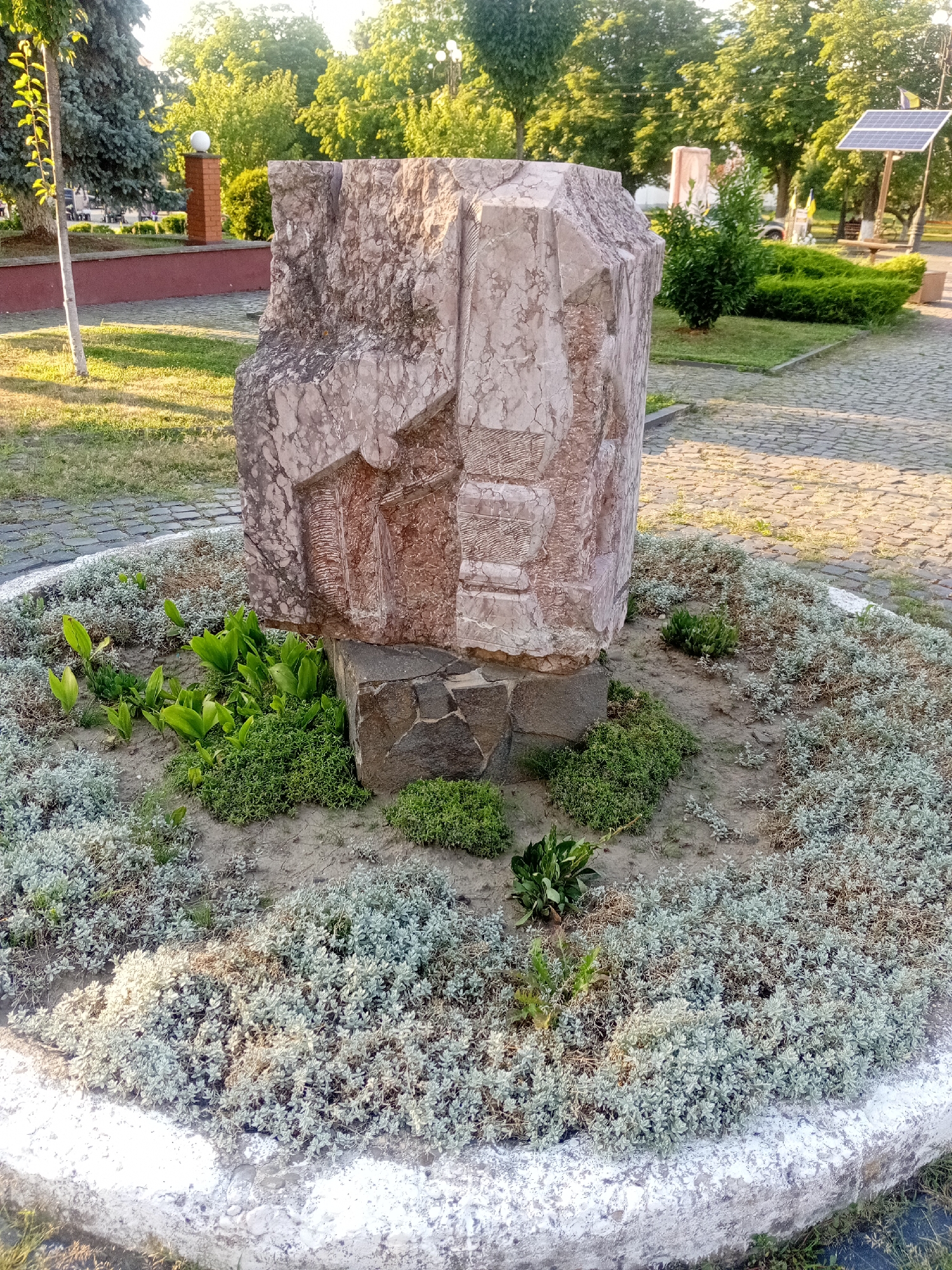 The image shows a monument in a park setting, likely in Tiachiv, Ukraine. The monument is made of large, rough-hewn stones, with the main piece appearing to be a light-colored, angular boulder with carved details or natural formations that resemble figures or symbols. It rests on a darker, smaller stone base. The monument is surrounded by a circular flower bed filled with low-lying, light green and greyish-green plants, with some taller, broader green leaves in the center. The flower bed is bordered by a white, concrete-like rim.
In the background, there is a paved pathway made of cobblestones, and further back, there are green trees and foliage. Sunlight casts shadows, suggesting it is daytime. To the right, a solar panel is visible on a wooden stand, with Ukrainian flags also present in the distance, reinforcing the location in Ukraine. Other elements in the background include a brick pillar with a spherical light fixture, a red retaining wall, and what appears to be a bench. No people are visible in the image. The overall scene depicts a monument in a public outdoor space, likely a park or square, maintained with landscaping.