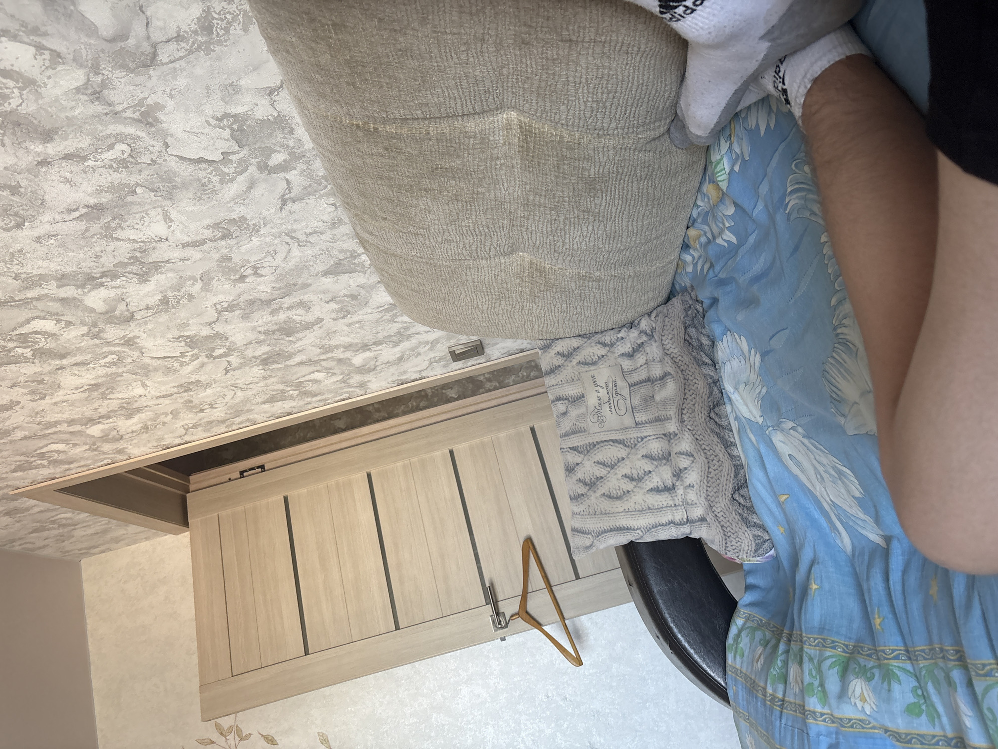 The image depicts an indoor scene, likely a bedroom or living area. A light-colored wooden door is partially open, revealing a darker interior. A wooden coat hanger hangs from the door handle. A textured wall with a marbled pattern covers the background. In the foreground, a beige upholstered sofa or armchair is visible, with a knitted pillow resting on it. A blue bedspread with a white bird and floral pattern covers a surface below the sofa. A person's legs, wearing white socks, are visible on the bedspread. The overall impression is one of a casual, domestic setting.
