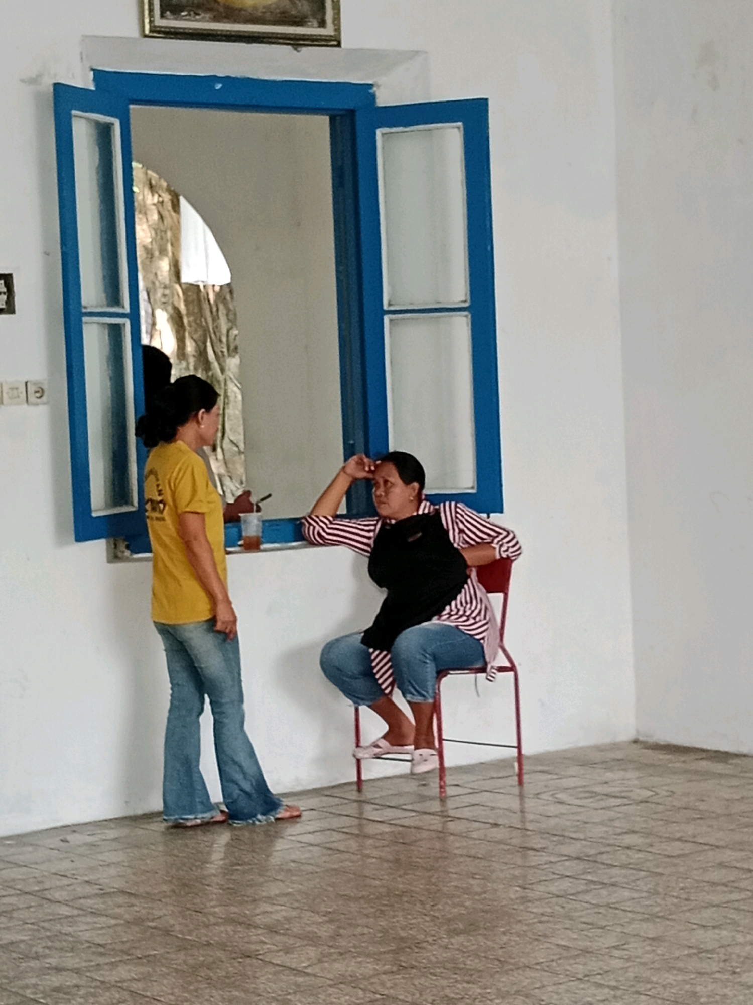 The image depicts an indoor scene with two women standing and sitting near a blue-framed window. One woman, wearing a yellow t-shirt and blue flared jeans, stands with her back to the viewer, facing a window. She has her hair pulled back in a bun. The other woman is seated on a red chair, wearing a striped shirt with red and white stripes and a black vest. She is looking towards the standing woman, with her hand resting on her forehead. A clear plastic cup with a brown liquid sits on the windowsill. The room has white walls and a tiled floor. The scene is set within a building, likely a public space given the open window and simple furnishings. The specific location in Jentekan, Indonesia, is not visually evident from the image itself.