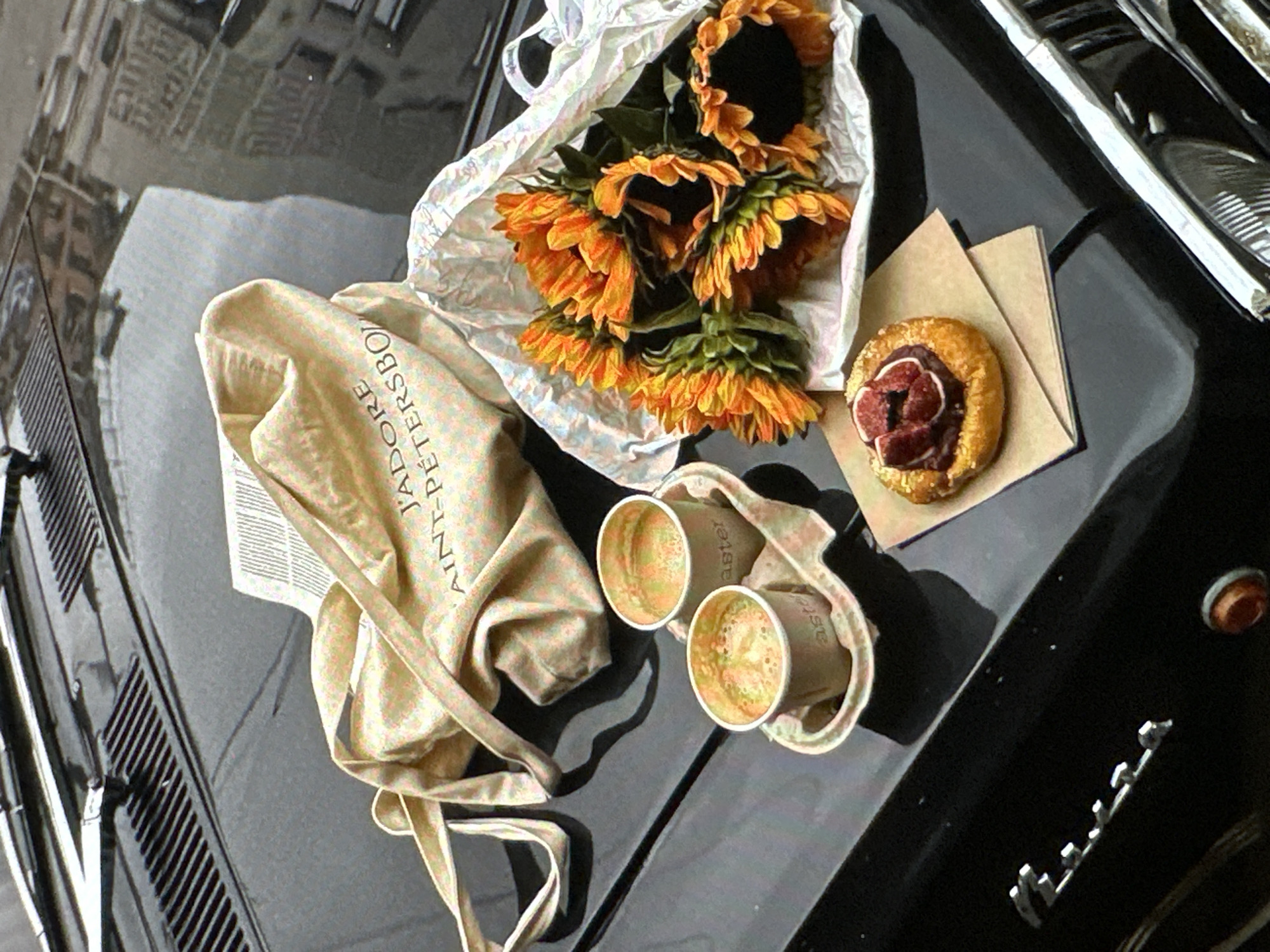 The image presents a still-life composition arranged on the hood of a black vintage car. The scene includes a bunch of sunflowers in a white plastic bag, a pastry with dark red fruit filling atop a folded brown paper napkin, two paper cups of coffee in a paper carrier, and a beige canvas tote bag with text printed on it. The location is suggested to be Yerevan, Armenia, based on the tote bag that reads "J'adore Saint-Petersbourg". The hood of the car reflects a blurry urban landscape, potentially indicating that the car is parked in a city setting. There are no people directly visible in the image.