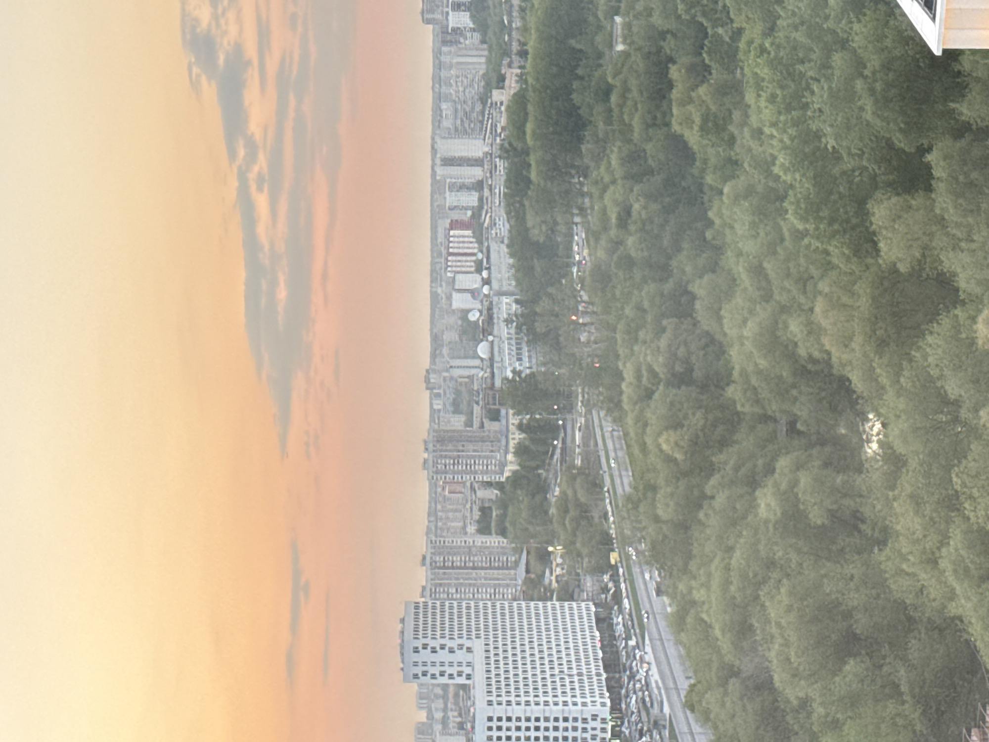 The image captures a cityscape at dusk or dawn, juxtaposing urban development with natural elements. A modern building with a white facade and grid-like windows occupies the lower left corner of the frame. In the middle, there are multiple buildings of different heights and sizes, also predominantly white or light-colored. To the right of the buildings, a dense green expanse of trees extends vertically, possibly forming a park or forest area. A road runs adjacent to the trees, lined with parked cars. The sky transitions from a bright, diffused light on the left to darker hues towards the upper portion of the frame. Clouds are visible, adding texture to the sky. The image suggests a location where urban and natural landscapes merge.