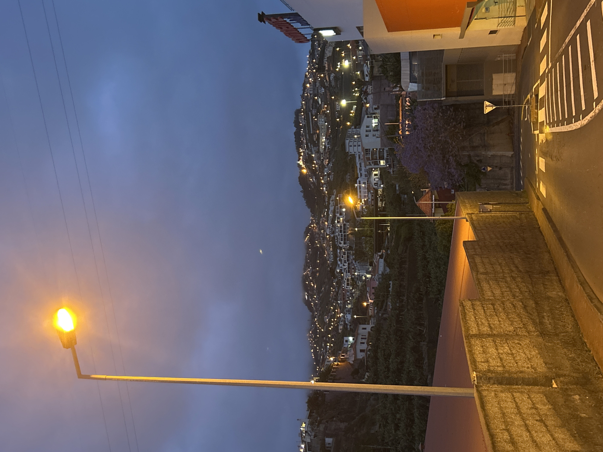 The image shows a nighttime view of Câmara de Lobos, Portugal, featuring a hillside densely populated with buildings and illuminated by numerous lights. A street lamp is in the foreground. Power lines extend across the sky. A street with a zebra crossing and sidewalk are visible on the right, with buildings on either side. The sky is a deep blue, suggesting early evening or late night. A faint crescent moon is visible.