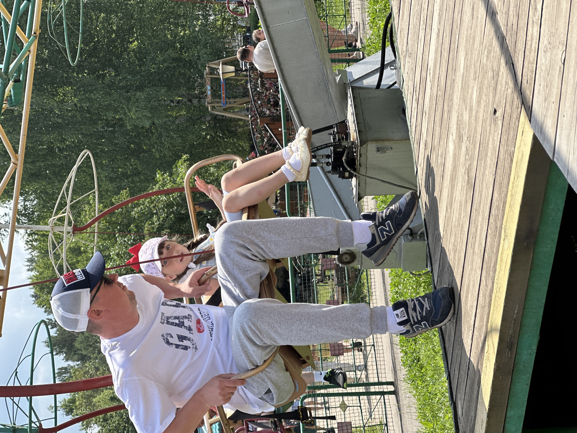 An adult male and a young girl are seated on a carousel-like ride at an amusement park. The male is wearing a white t-shirt, grey sweatpants, and a blue and white baseball cap. He is looking down and to his right. The girl is wearing a light-colored dress and a pink hat with a red bow. She is looking forward with her mouth slightly open. They are both holding onto a wooden bar. The ride appears to be made of metal and wood. In the background, there are trees and other parts of the amusement park. The ground is made of wooden planks. The location is identified as an amusement park in Chaikovsky, Russia.