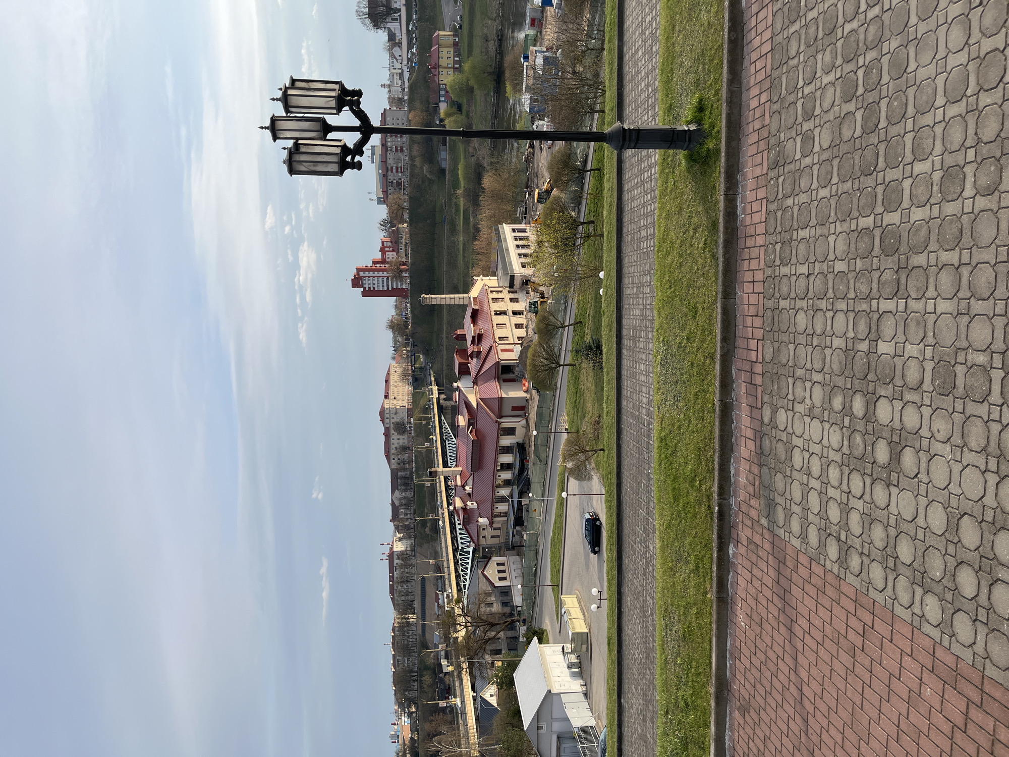 The image captures a cityscape viewed from an elevated position, possibly a walkway or embankment. In the foreground, a black street lamp with three lanterns stands tall on the right side of the frame, with a section of patterned paving stones and a strip of green grass immediately adjacent to it.
The middle ground reveals a more distant urban landscape. There are various buildings, including structures with red roofs and others with more modern architecture. A bridge with a distinctive lattice structure spans across what appears to be a waterway or a lower street. The presence of trees and green spaces interspersed between the buildings suggests a partially landscaped environment. A dark-colored car is visible on the road.
The sky above is partly cloudy, with areas of blue visible between the clouds, indicating fair weather. The composition of the image suggests it was taken during daylight hours, with the light casting shadows that provide depth to the scene.