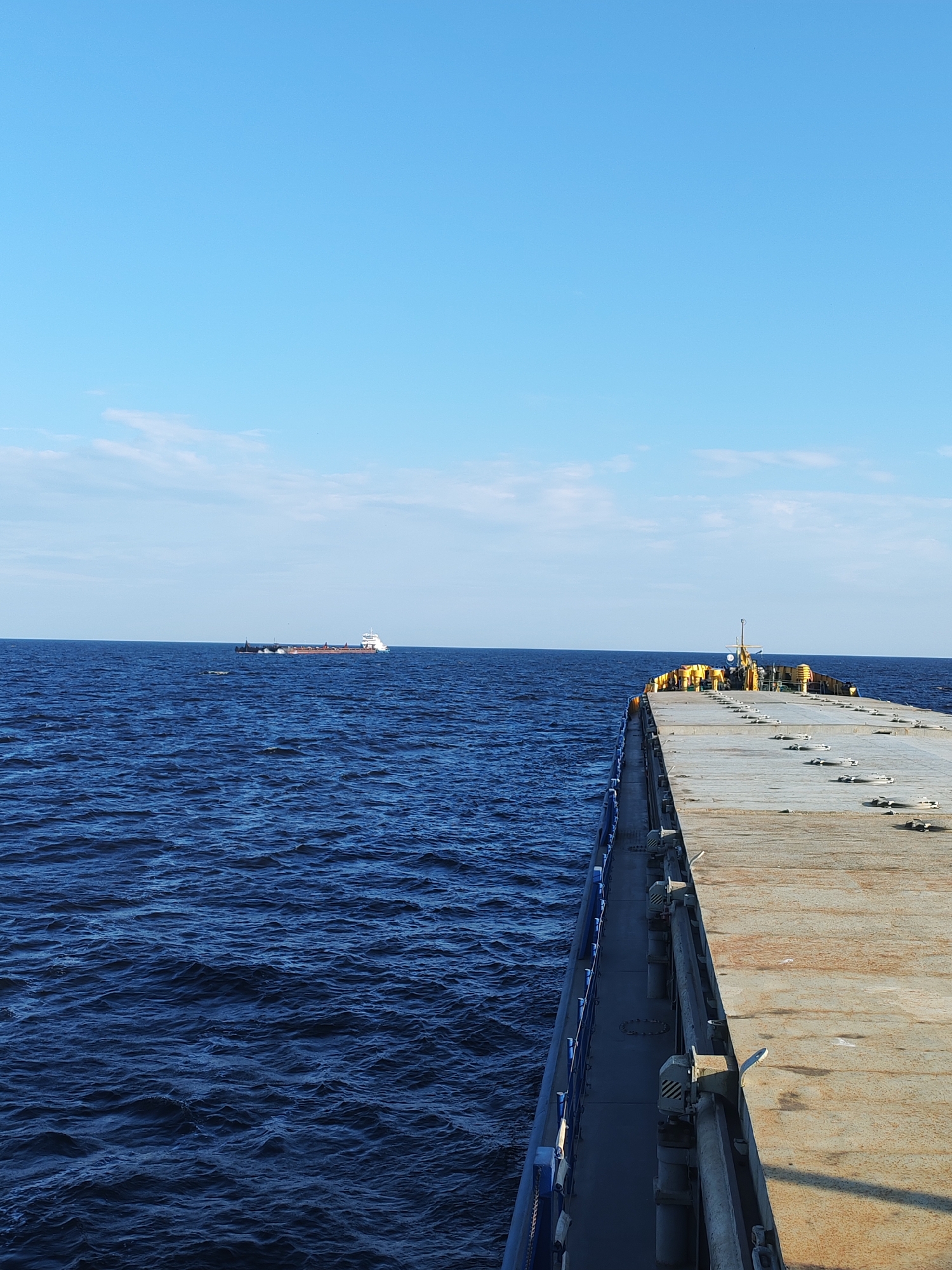 This media file captures a wide-angle view of a vessel at sea under a clear, bright sky. The main subject is a large, flat-decked cargo ship, its deck appearing weathered and utilitarian, with numerous tie-down points visible. The ship is on the right side of the frame, extending into the distance. To the left, the deep blue, slightly rippled ocean stretches to the horizon. In the distance, a white and red cargo barge is visible, also on the water. The scene suggests a bright, sunny day with a clear sky, likely late morning or early afternoon given the lighting. The overall impression is one of maritime transport and open sea. There is no visible text in the image.