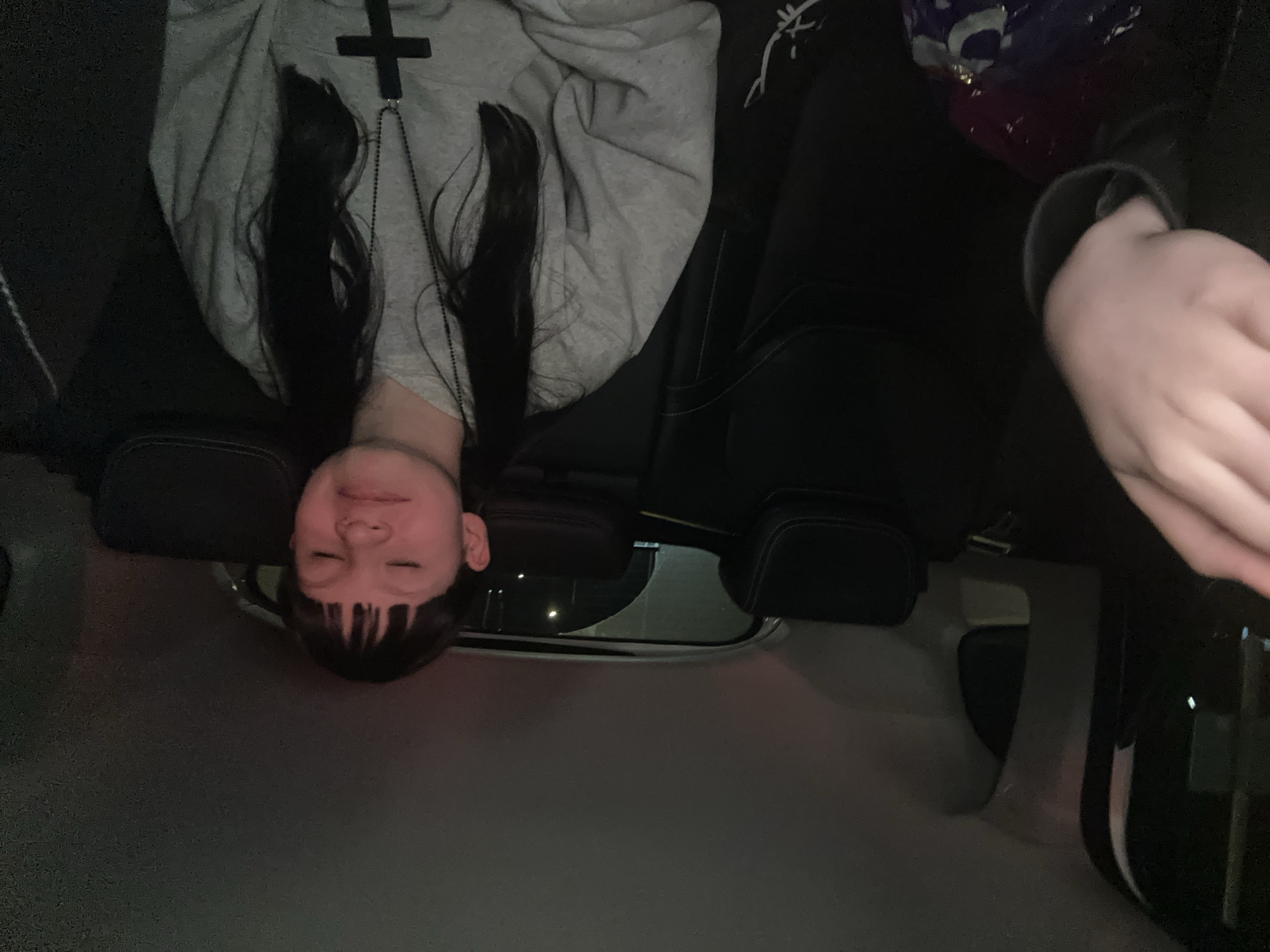 A photograph depicts a young person lying down in what appears to be the back seat of a car. The person has dark hair with bangs and is wearing a gray sweatshirt with a black cross design. Their eyes are closed. The car's interior includes black leather seats and dark trim. The location cannot be determined from the image.