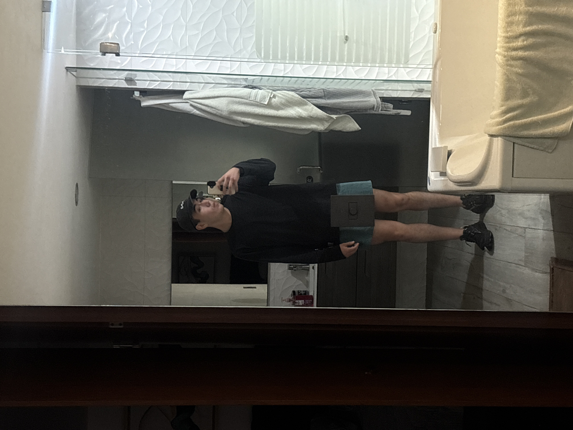 A man in a black long-sleeved shirt, blue shorts, and a baseball cap is taking a picture of himself in a bathroom mirror. The bathroom has white textured wall tiles, a glass shower enclosure, a toilet with a towel on the tank, and a vanity with drawers. There is no explicit evidence in the image to confirm that the location is San Bernardino Tlaxcalancingo, Mexico.