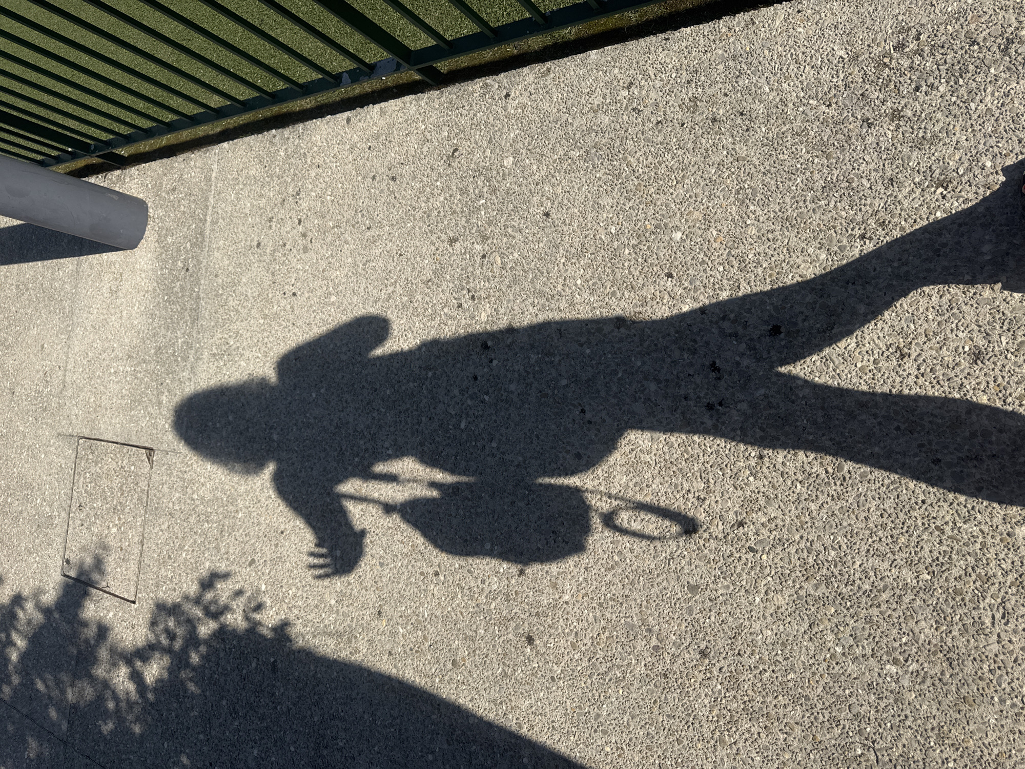 The image shows the shadow of a person on a concrete path in Saint-Claude, France. The person is casting a shadow that is shaped like a human form, with a backpack slung over one shoulder. The shadow appears to be reaching out with one hand. The concrete path is textured and speckled with small pebbles. To the right of the path, there is a green area, possibly a sports field, bordered by a dark green metal fence. A grey cylindrical pole casts a shadow on the path from the upper right side of the image. In the background, a faint shadow of tree leaves can be seen on the concrete. The lighting suggests it is daytime, with the sun casting strong shadows.