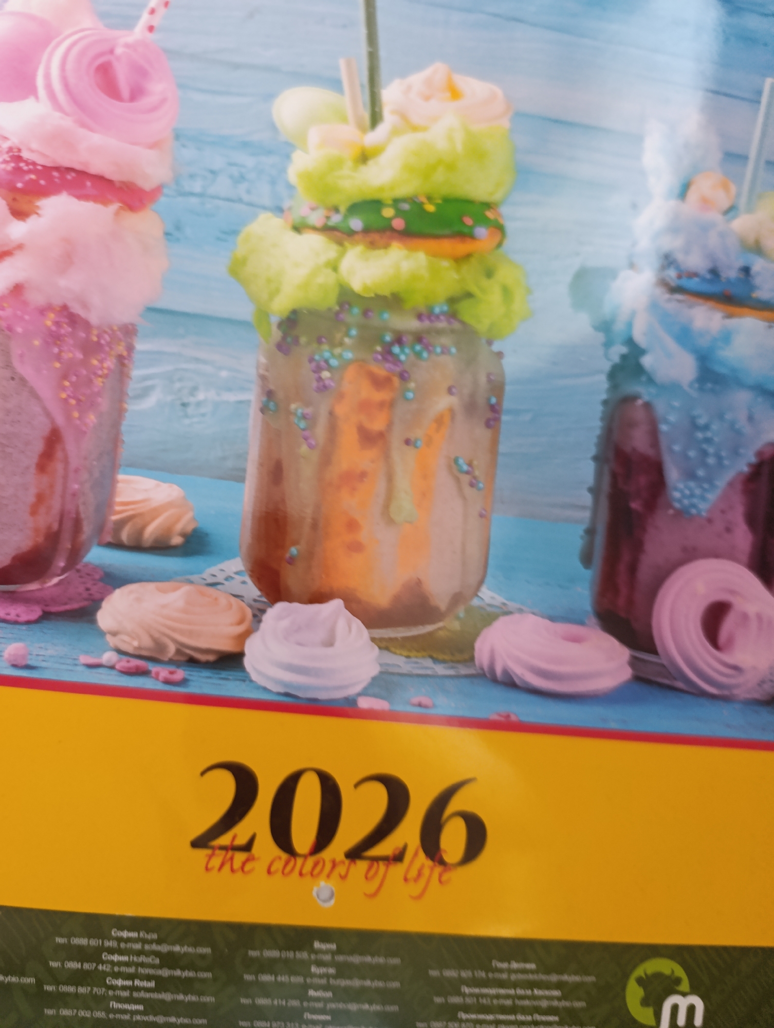 The image is a promotional photograph featuring three colorful, elaborately decorated milkshakes or ice cream sundaes served in large glass jars. The desserts are stacked high with whipped cream, cotton candy, cookies, and sprinkles, creating a whimsical and decadent presentation. In the foreground, several meringue cookies in pastel colors are scattered on a blue wooden surface.
At the bottom of the image, a yellow band displays the text "2026 the colors of life" in large, dark font. Below this, a dark green band contains contact information for various locations.
Specifically, the contact information for the city of Razgrad, Bulgaria is:
**Разград**
тел.: 0888 018 505, e-mail: razgrad@miky.bio.com