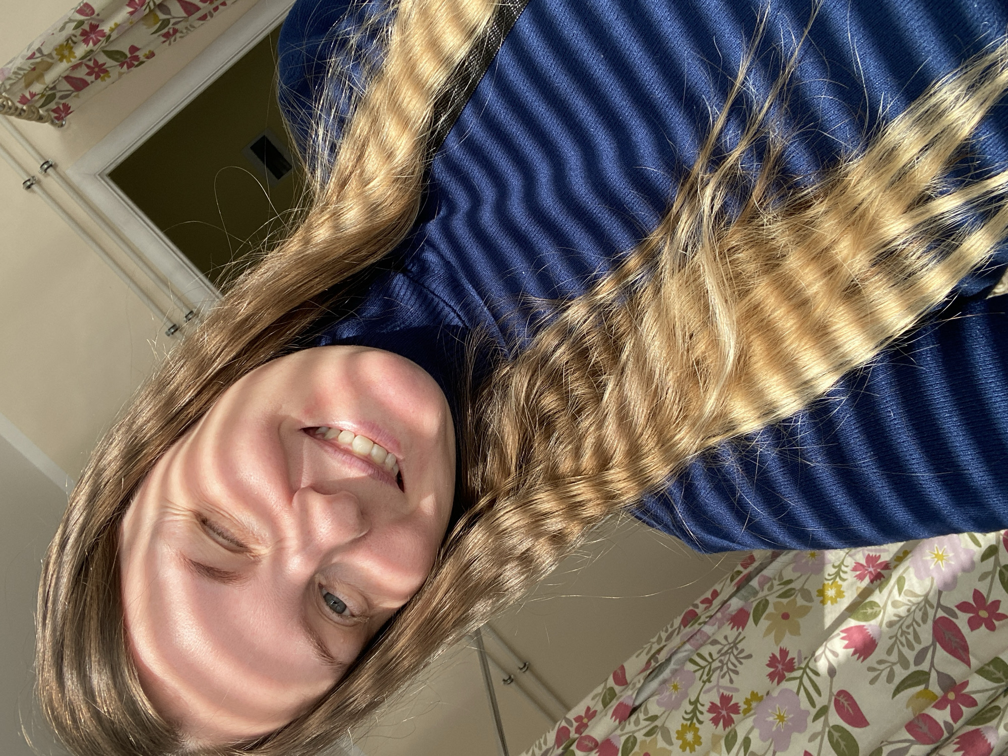 A close-up, upward-angled shot in Noviy, Russia, captures a young woman smiling broadly with her eyes squinting slightly, illuminated by bright sunlight. Her long, light brown hair falls around her face and extends across the frame. Distinct striped shadows, likely cast by window blinds, stretch across her forehead, cheek, nose, and lips, as well as over her hair and the dark blue, ribbed sweater she is wearing. The sweater's texture is clearly visible through the shadows. In the upper left corner, a white window frame is partially visible, offering a glimpse into a darker interior. The lower right portion of the image shows a fabric with a cheerful floral pattern featuring pink, green, and yellow flowers on a light background, possibly curtains or bedding. The bright, directional sunlight suggests it is daytime. The woman appears happy and relaxed. There is no visible text.