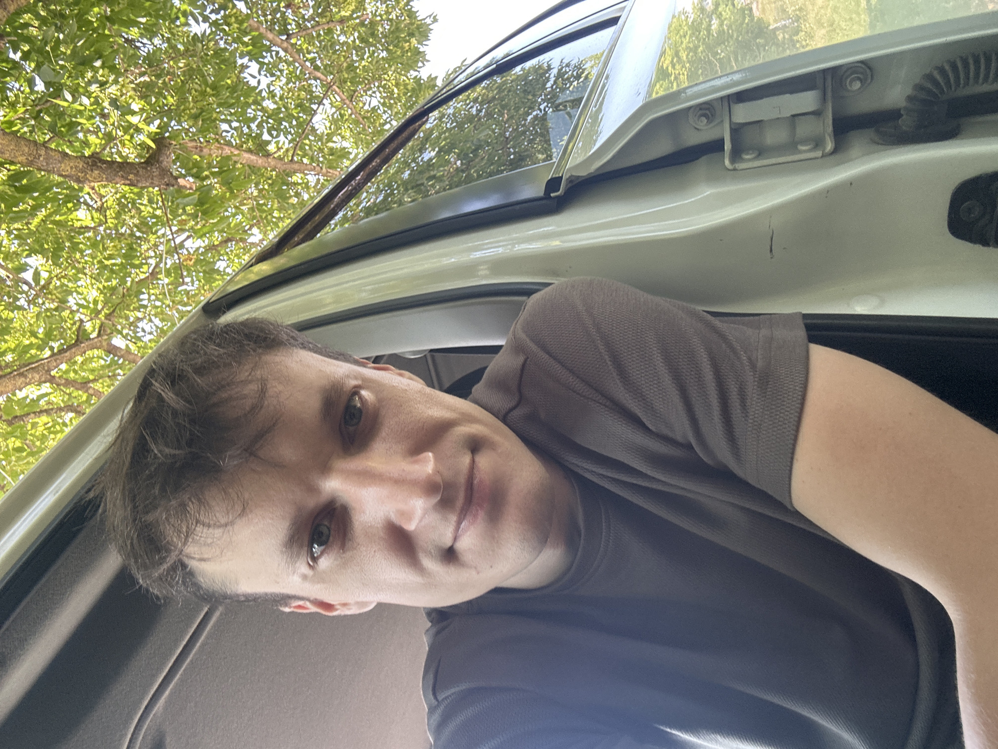 A man is taking a selfie inside a car. The man is positioned in the driver's seat, with the car door open. He is looking directly at the camera with a slight smile. His left arm is resting on the open car door. The background of the image shows green foliage and trees, suggesting the car is parked outdoors in a natural setting. The car appears to be light-colored, possibly white or cream. The lighting indicates it is daytime, with sunlight filtering through the leaves. The scene does not provide any specific indicators of being in the city of Makiv, Ukraine.